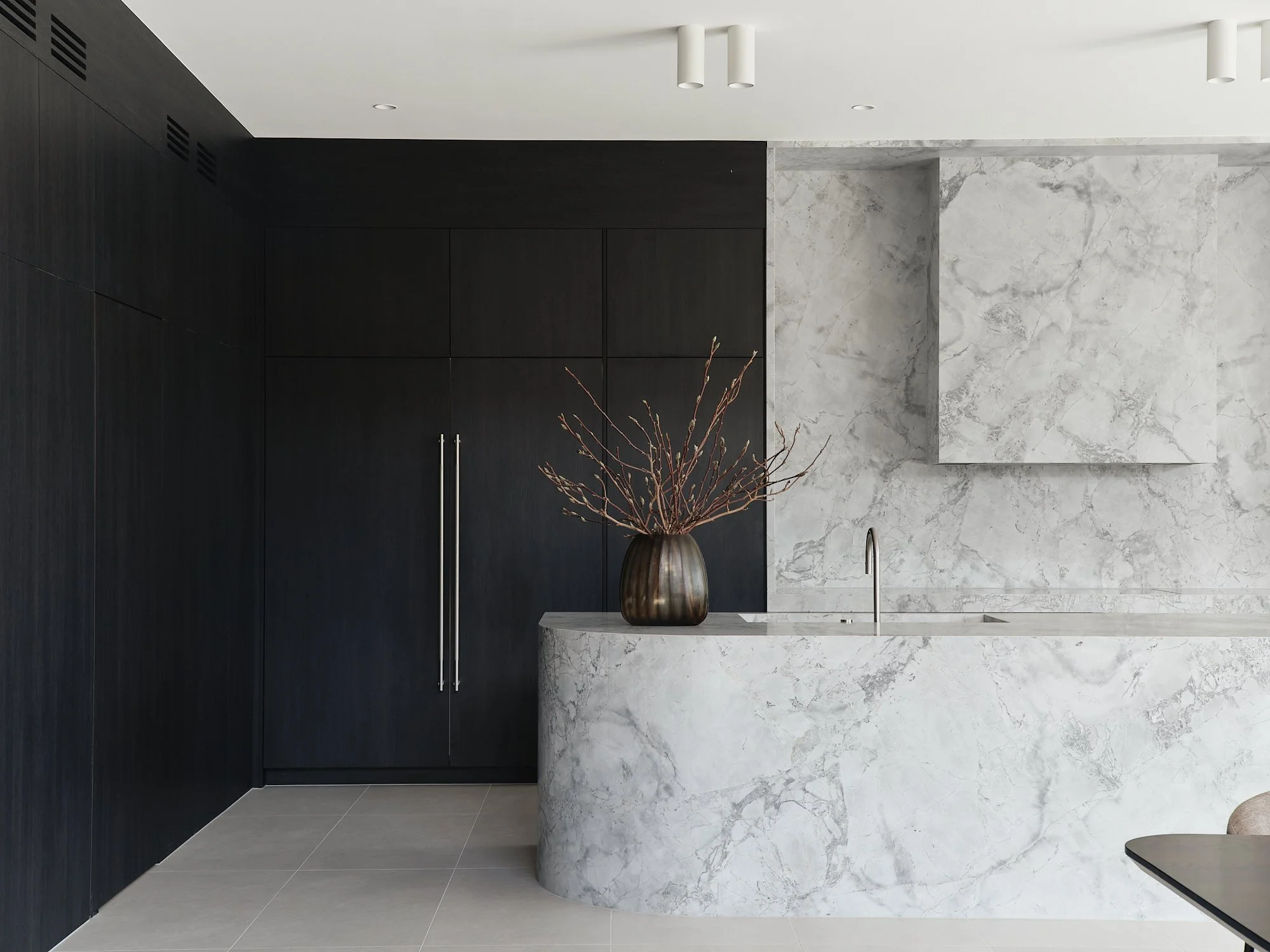 Dark kitchen cabinetry beside a pale veined stone island and matching stone splashback.