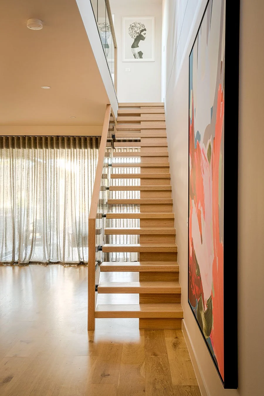 Interior view of a modern house with a wooden staircase, colorful abstract wall art on the right, a framed black-and-white silhouette portrait of a person with curly hair at the top of the stairs, and a large window with curtains on the left.