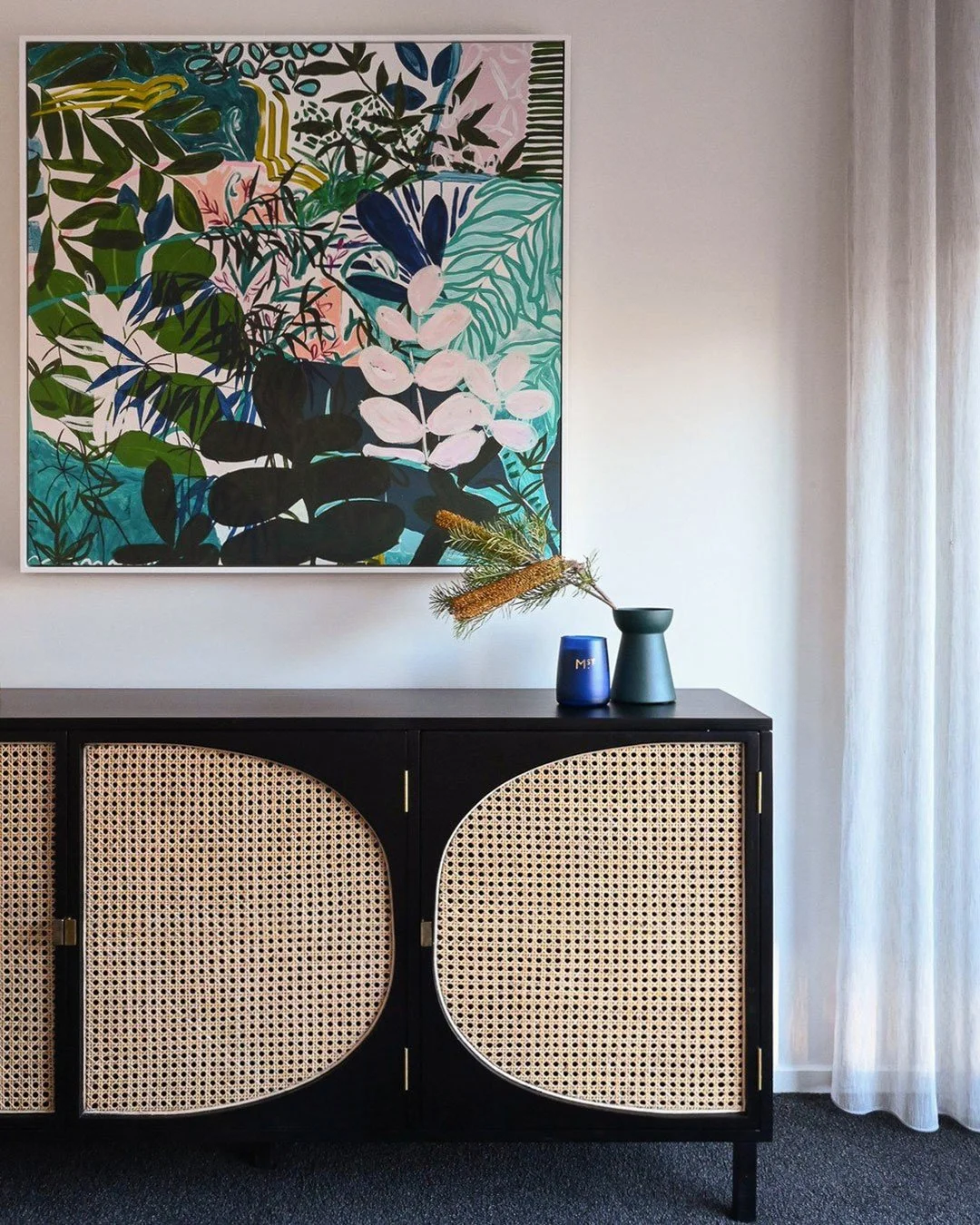 A black sideboard with rattan double doors, a large framed botanical art print on the wall above, and a blue vase with dried flowers on top, positioned in a room with white walls and curtains.