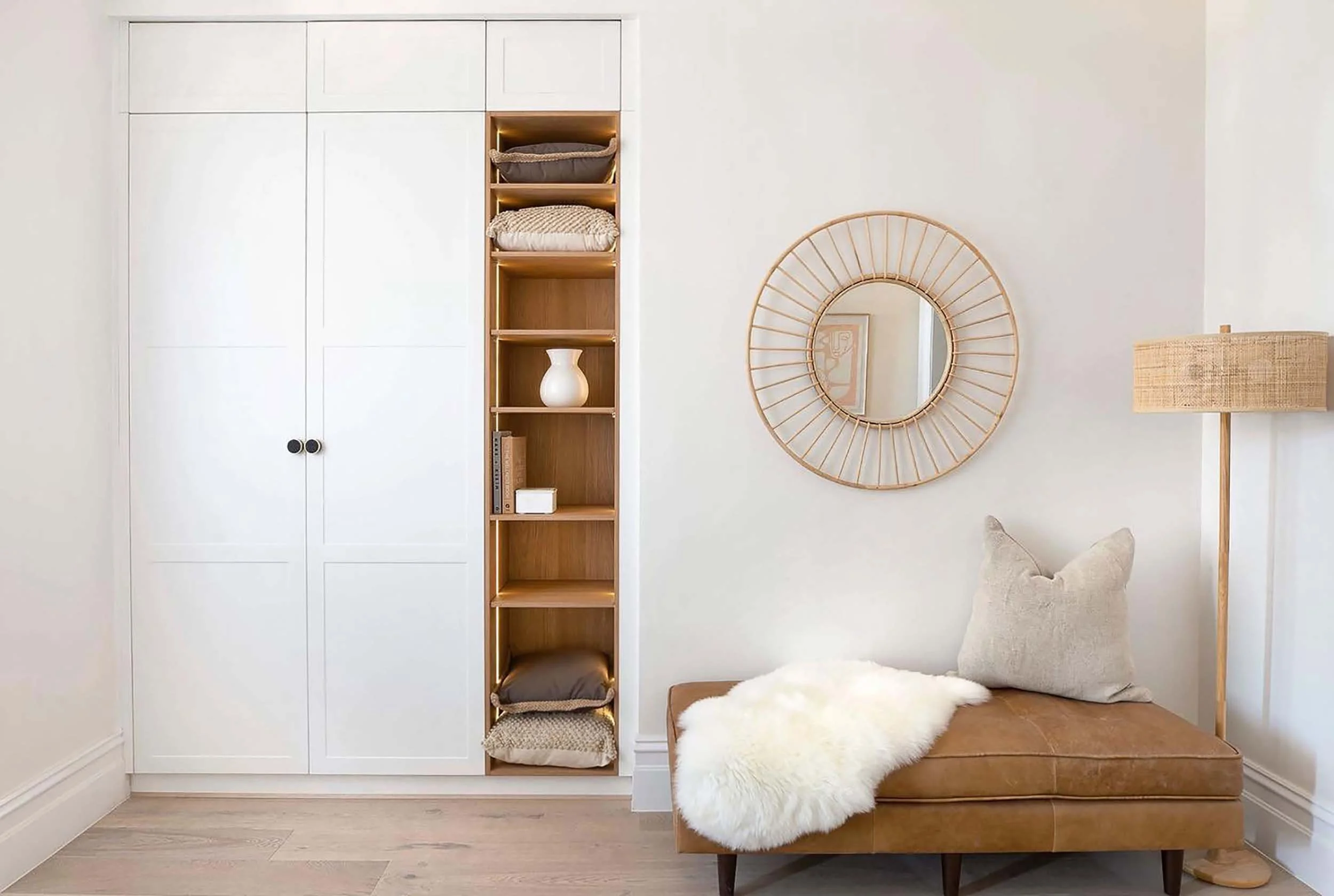 Interior of a minimalist room with a white built-in wardrobe, a wooden open shelf with pillows and decorative items, a round mirror with a wooden frame, a beige floor lamp, and a brown leather bench with a white sheepskin throw and a beige pillow.