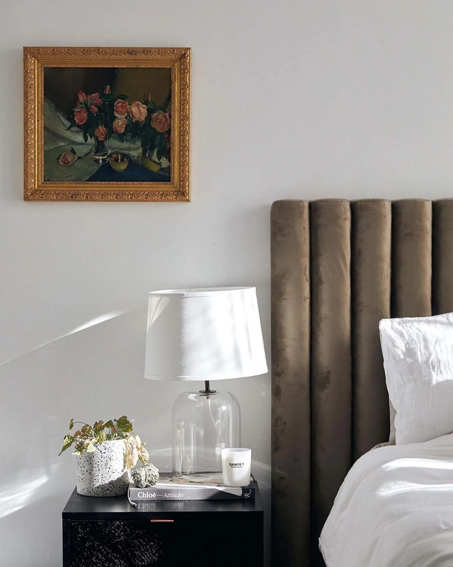 A bedroom corner with a beige upholstered headboard, a white bedside lamp, a black nightstand with decorative books, a candle, and a potted plant. A framed floral painting hangs on the white wall.