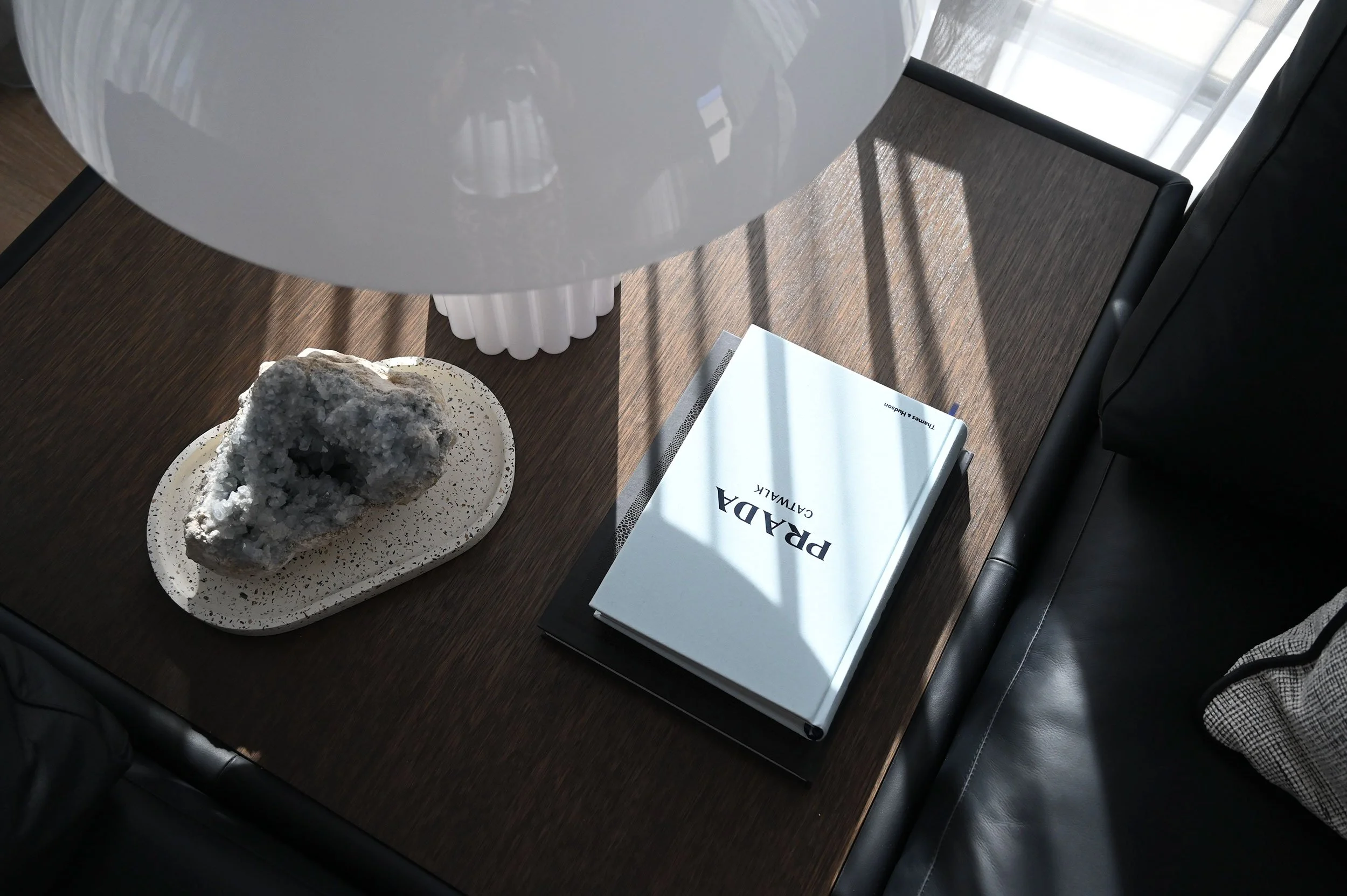 A wooden table with a large white spherical lamp, a decorative rock on a white plate, a book titled 'PRADA CALWAK,' and a closed notebook or journal.