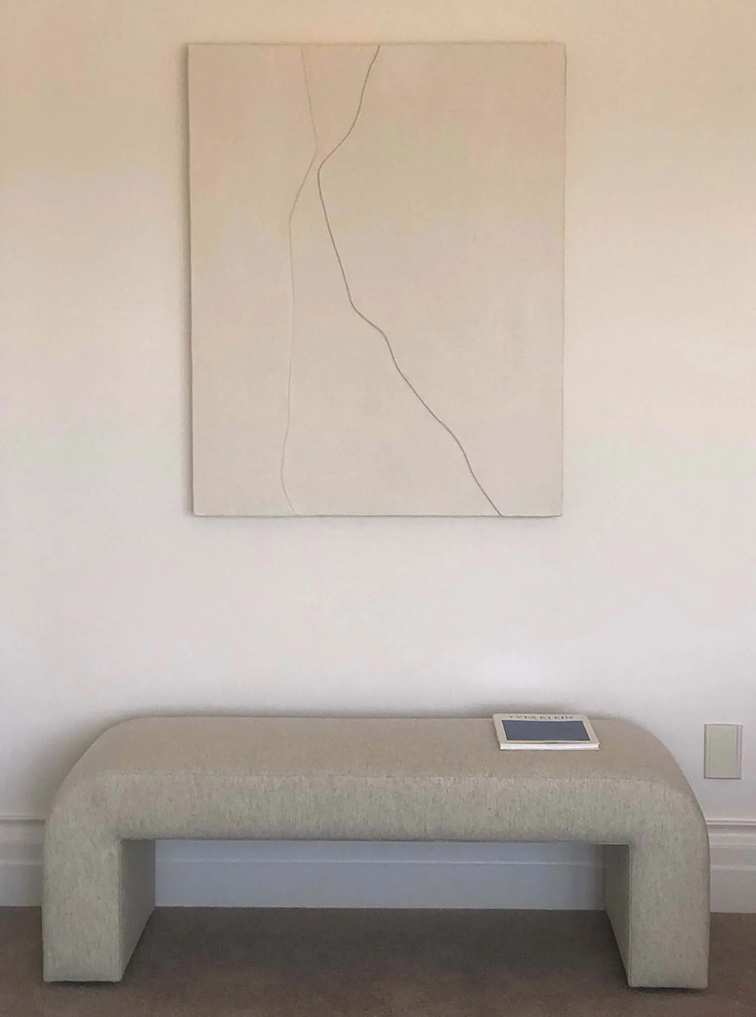 A blank white wall with a minimalist line drawing of a human face and neck, hanging above a beige couch in a gallery or museum setting, with a small digital display on the armrest.