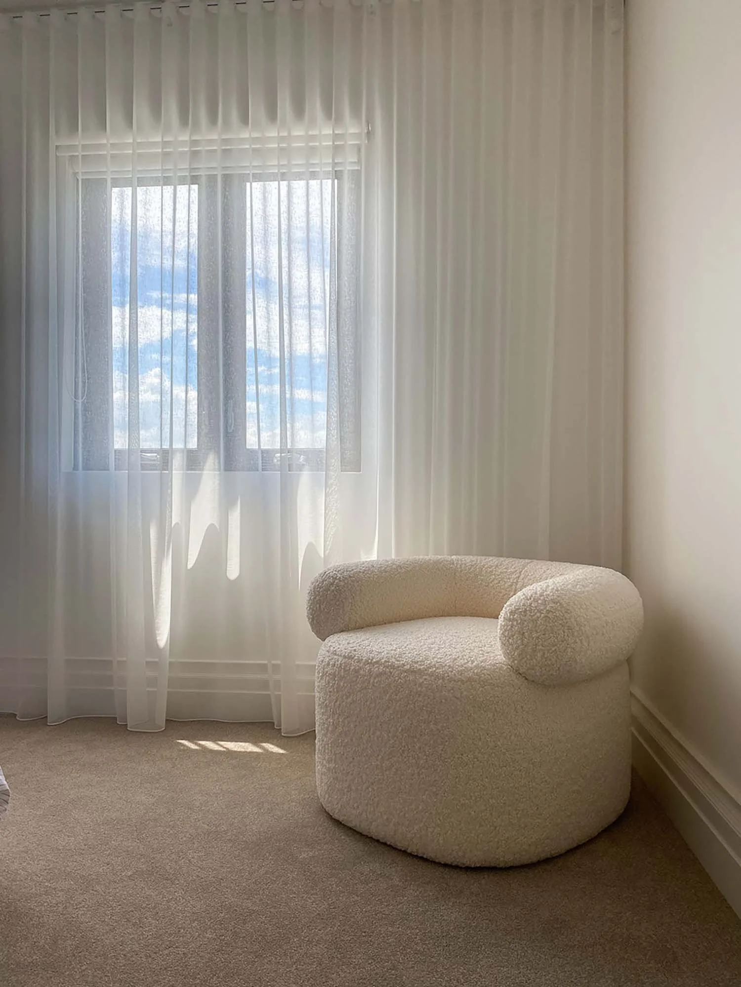 A beige, textured, rounded armchair is placed against a wall with a window covered by sheer white curtains. The window reveals a blue sky with clouds outside. The room has beige carpeting and white walls.