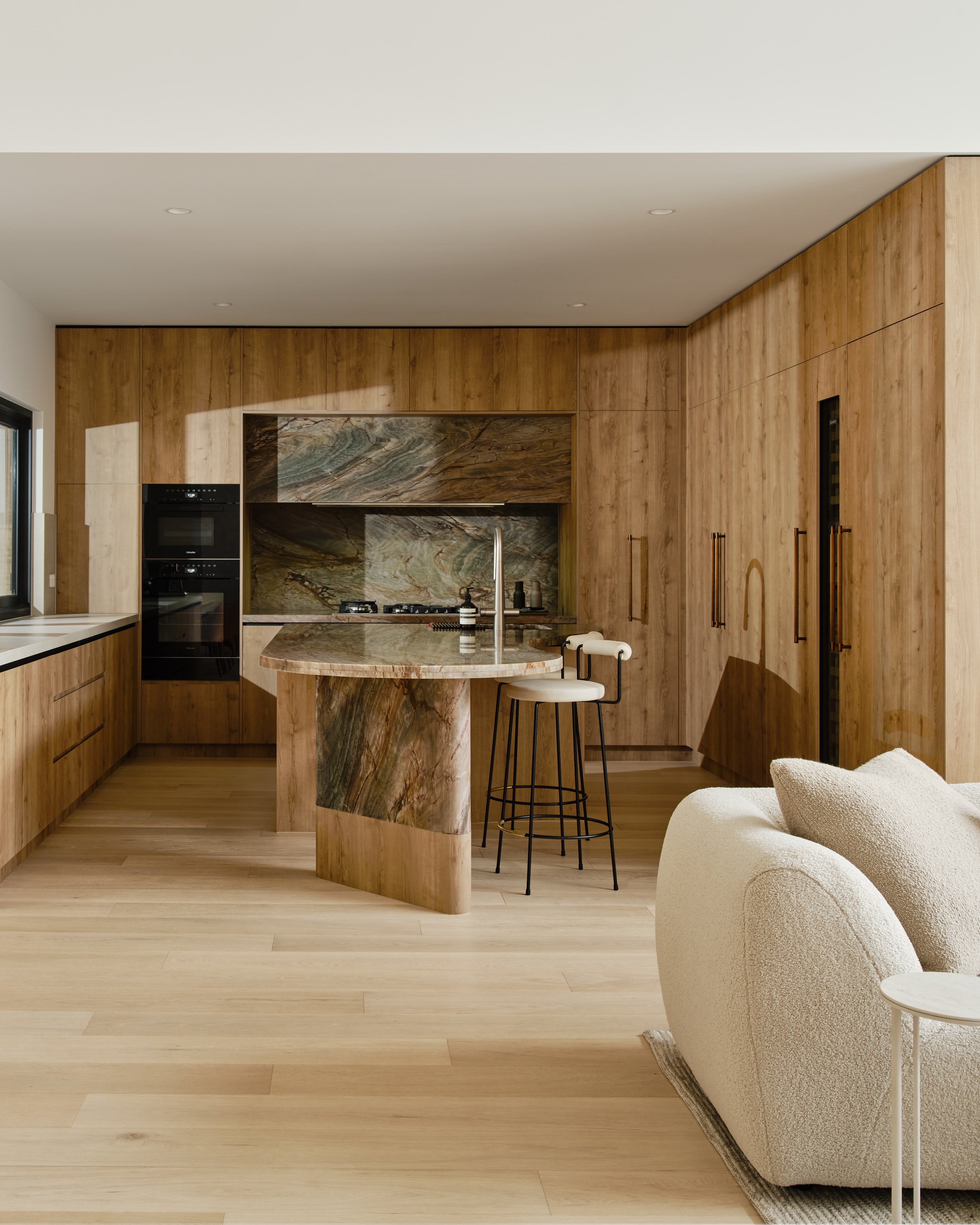Kitchen and adjoining living area with warm timber cabinetry, stone surfaces and soft upholstered seating.