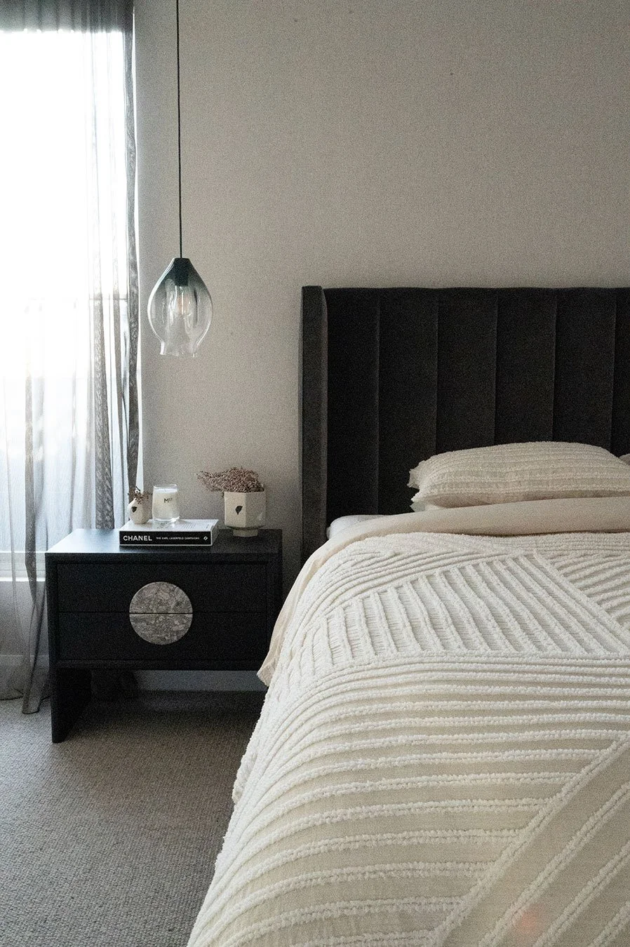 A bedroom with a bed, black headboard, cream textured blanket, and a nightstand with a book titled 'Chanel', a candle, a decorative bowl, and a small container. A pendant light hangs next to a window with sheer curtains.
