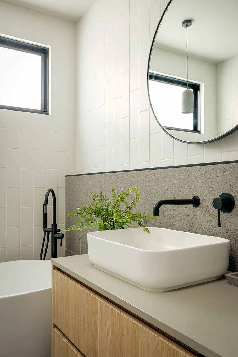 Modern bathroom with a white vessel sink, black fixtures, a large round mirror, a window, a pendant light, and green plant on the counter.