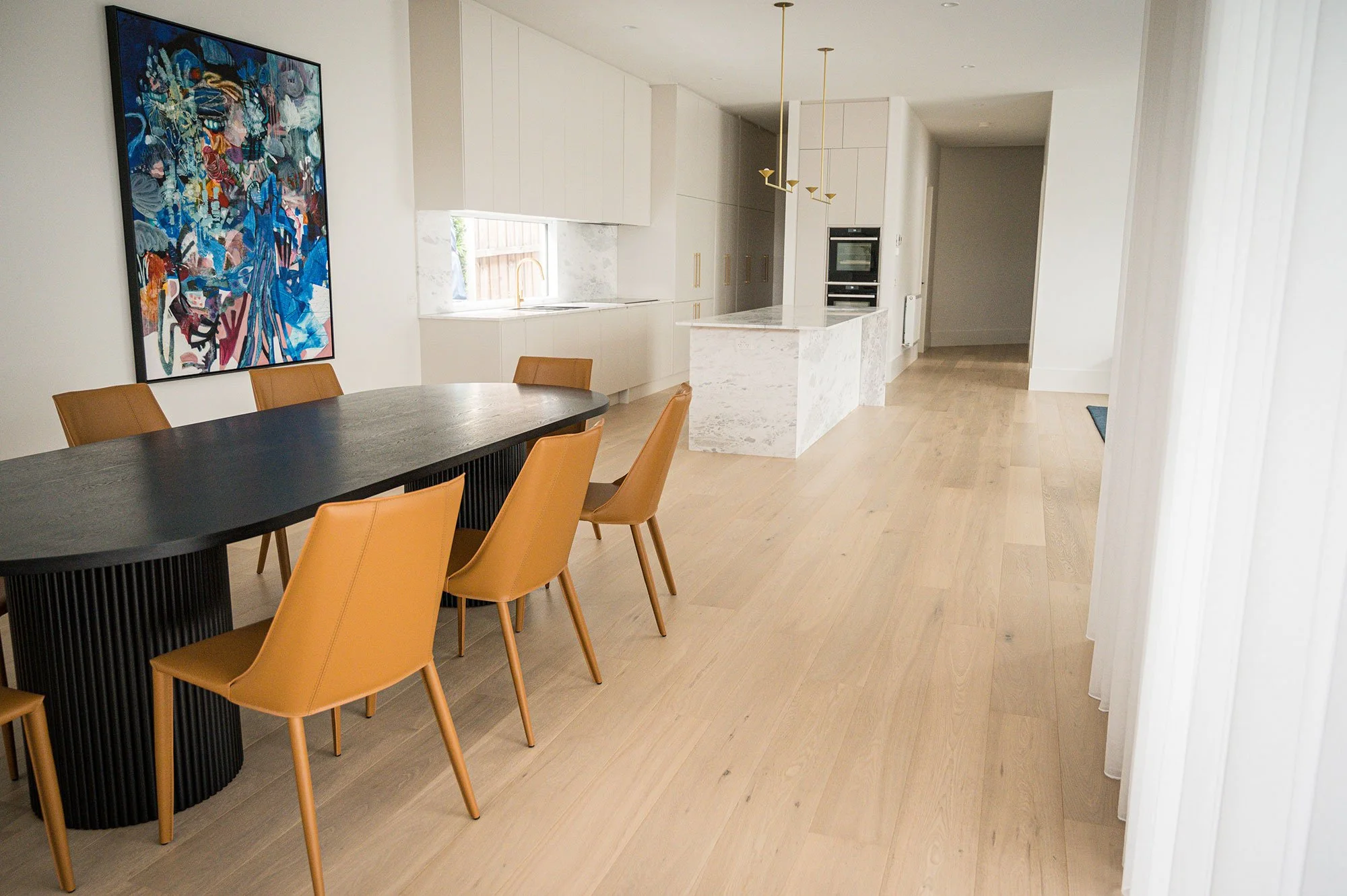 Modern kitchen and dining area with light wood flooring, white cabinets, marble island, a black dining table with tan chairs, and a colorful abstract painting on the wall.