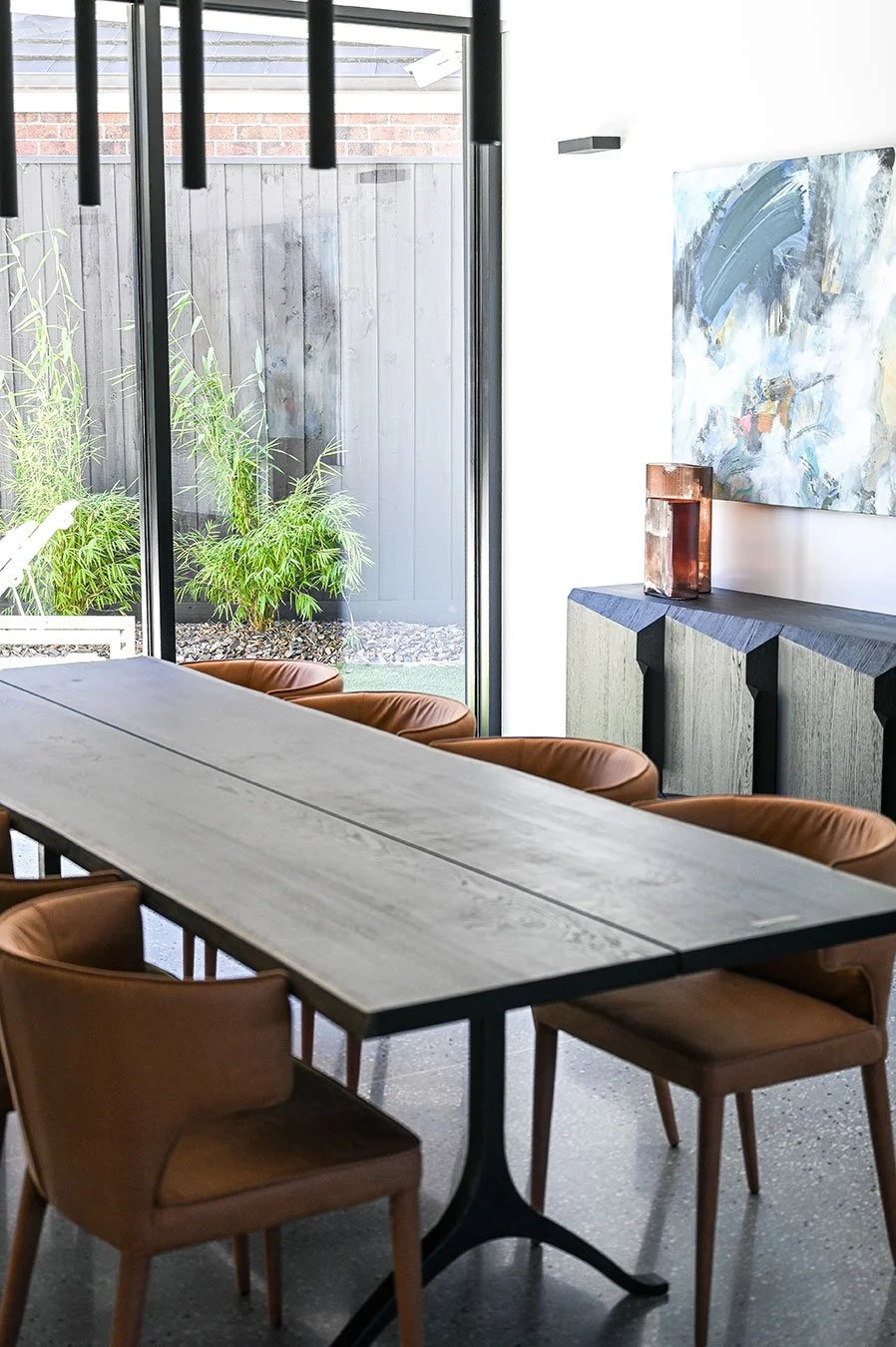 Modern dining area with a large wooden table surrounded by leather chairs, large glass sliding door with greenery outside, and abstract wall art with a cabinet and decorative object inside.