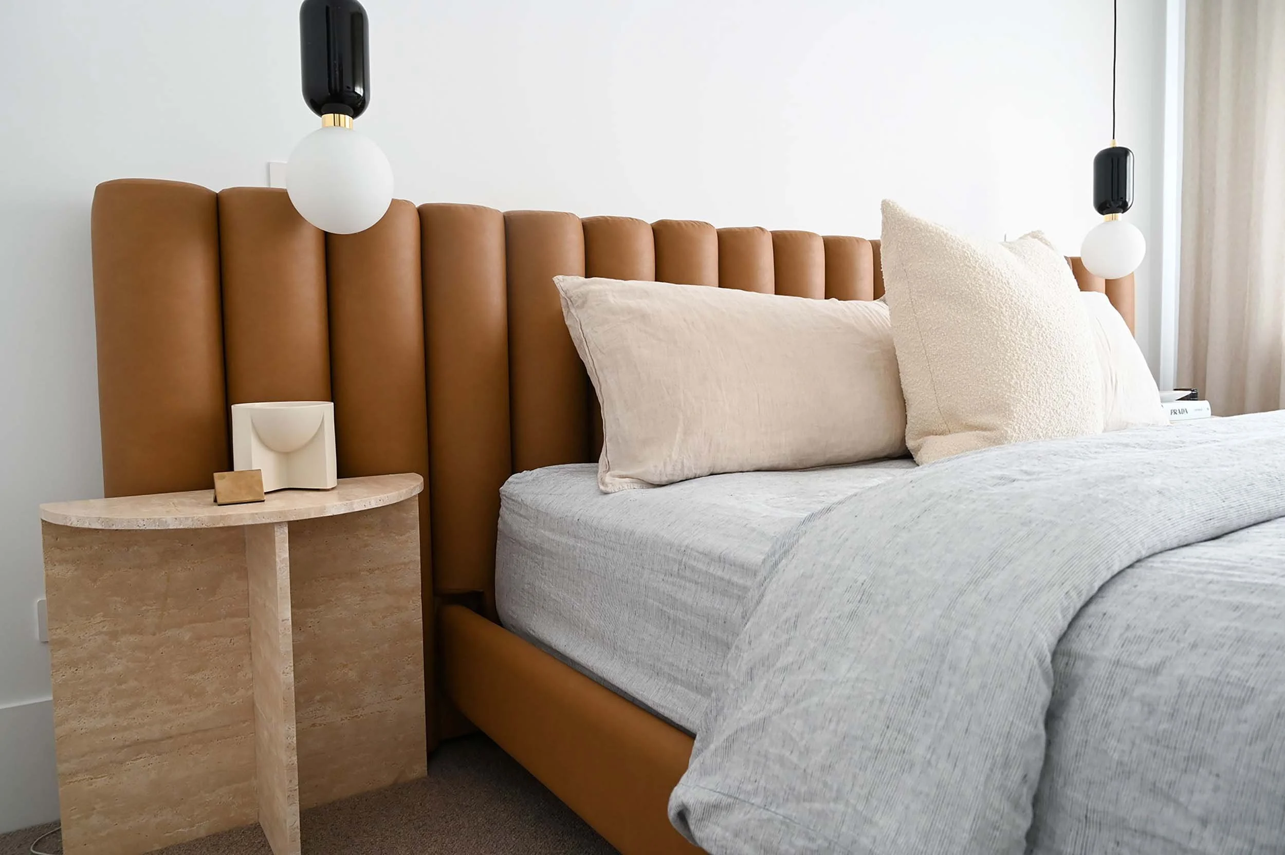 Close-up of a bedroom with a tan leather headboard, beige pillows, a beige side table, and modern hanging black and white pendant lights.