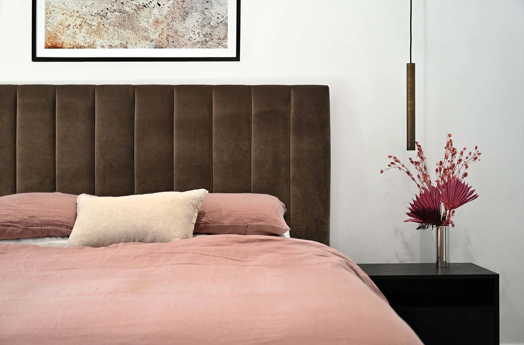 A modern bedroom with a brown upholstered headboard, pink bedspread, and matching pillows. To the right, a black nightstand holds a tall glass vase with pink and red flowers, and a vertical hanging light fixture is beside it. A framed abstract artwor