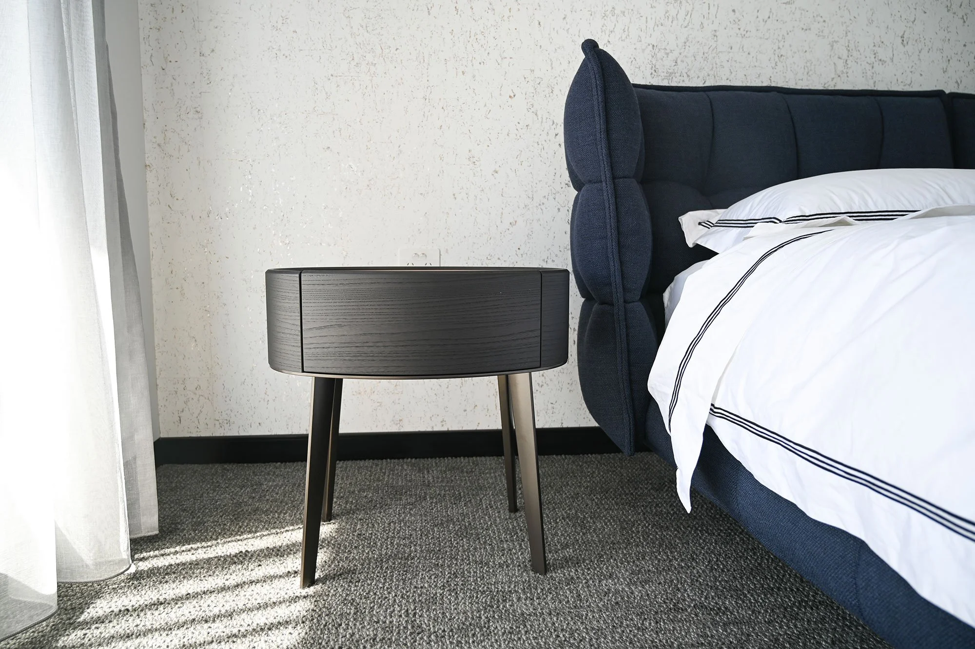 Side view of a modern bedroom featuring a dark blue upholstered bed with white and black striped bedding, a black wooden bedside table, a textured white wall, gray curtains, and gray carpeted flooring.
