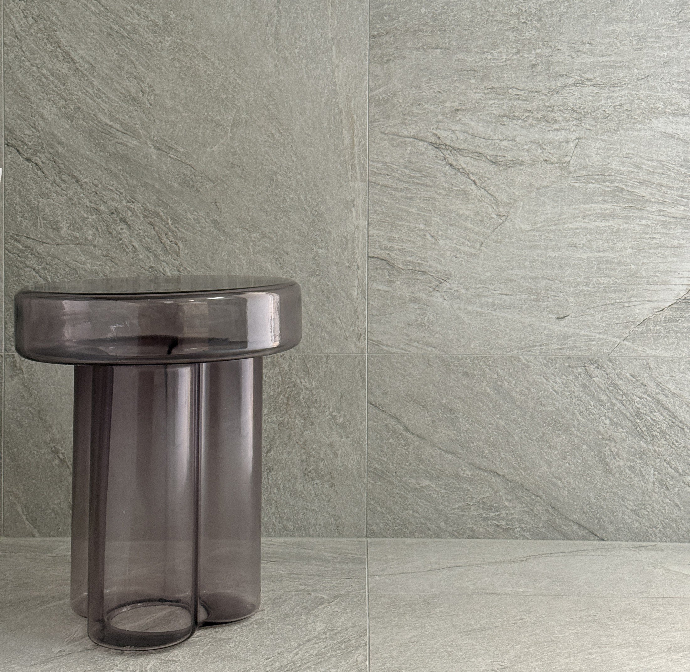 Translucent grey sculptural stool against pale stone bathroom tiles.