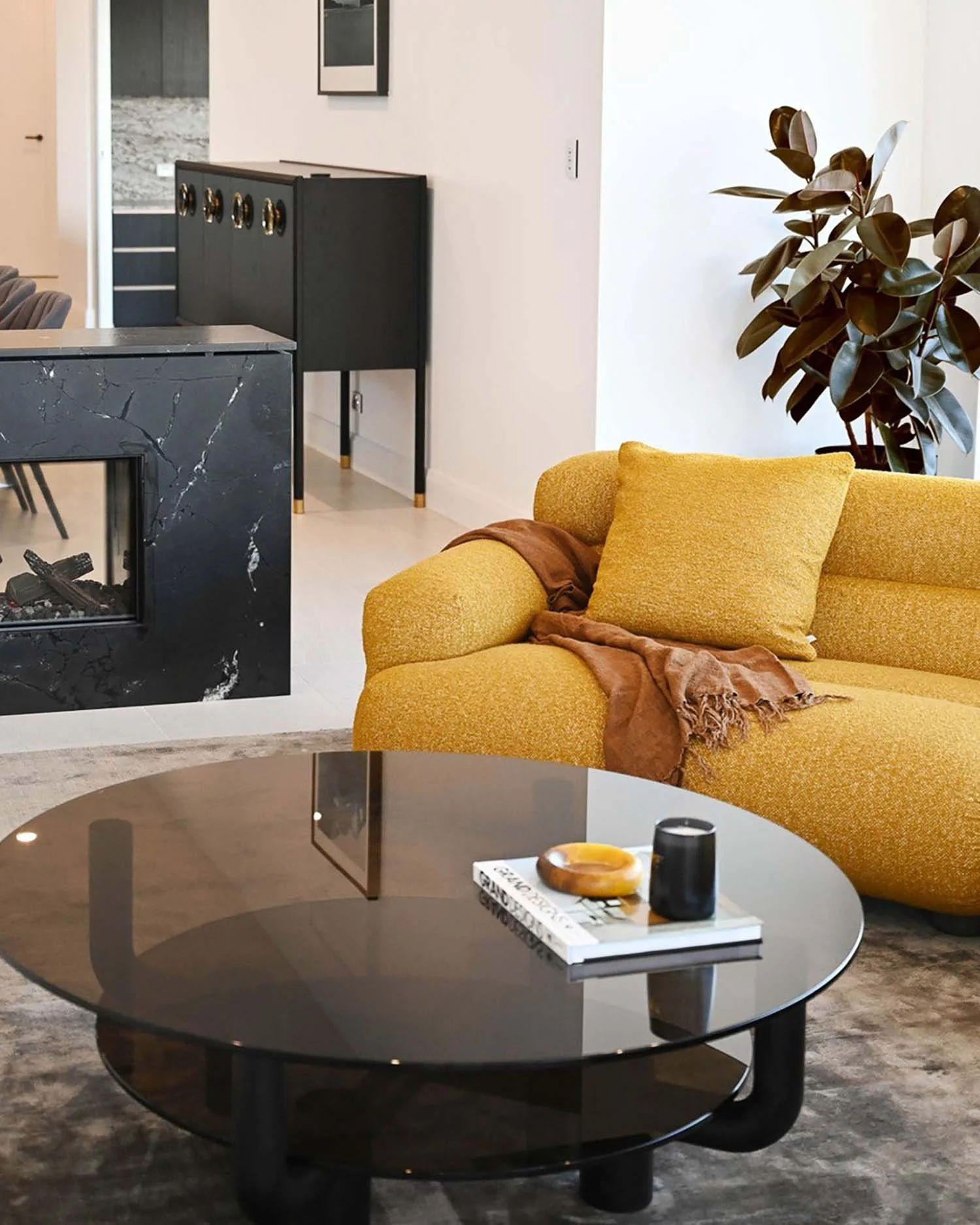 Living room with a yellow sofa, a dark round coffee table with a book, a yellow ceramic bowl, and a black cylindrical container, a green plant, and a black fireplace, with a kitchen area visible in the background.