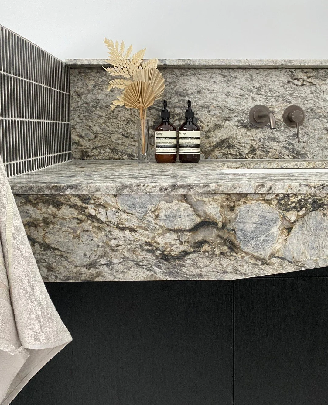 Close-up of a modern bathroom sink with a marbled countertop, a faucet, two soap dispensers, and a decorative vase with dried leaves and fan palm leaves.