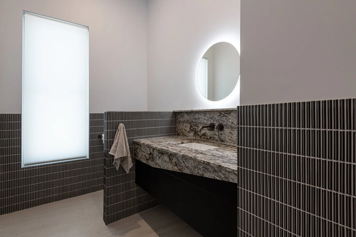 Modern bathroom featuring a frosted window, black tiled walls, a marble vanity with a sink, a round backlit mirror, and a beige towel hanging on a wall hook.
