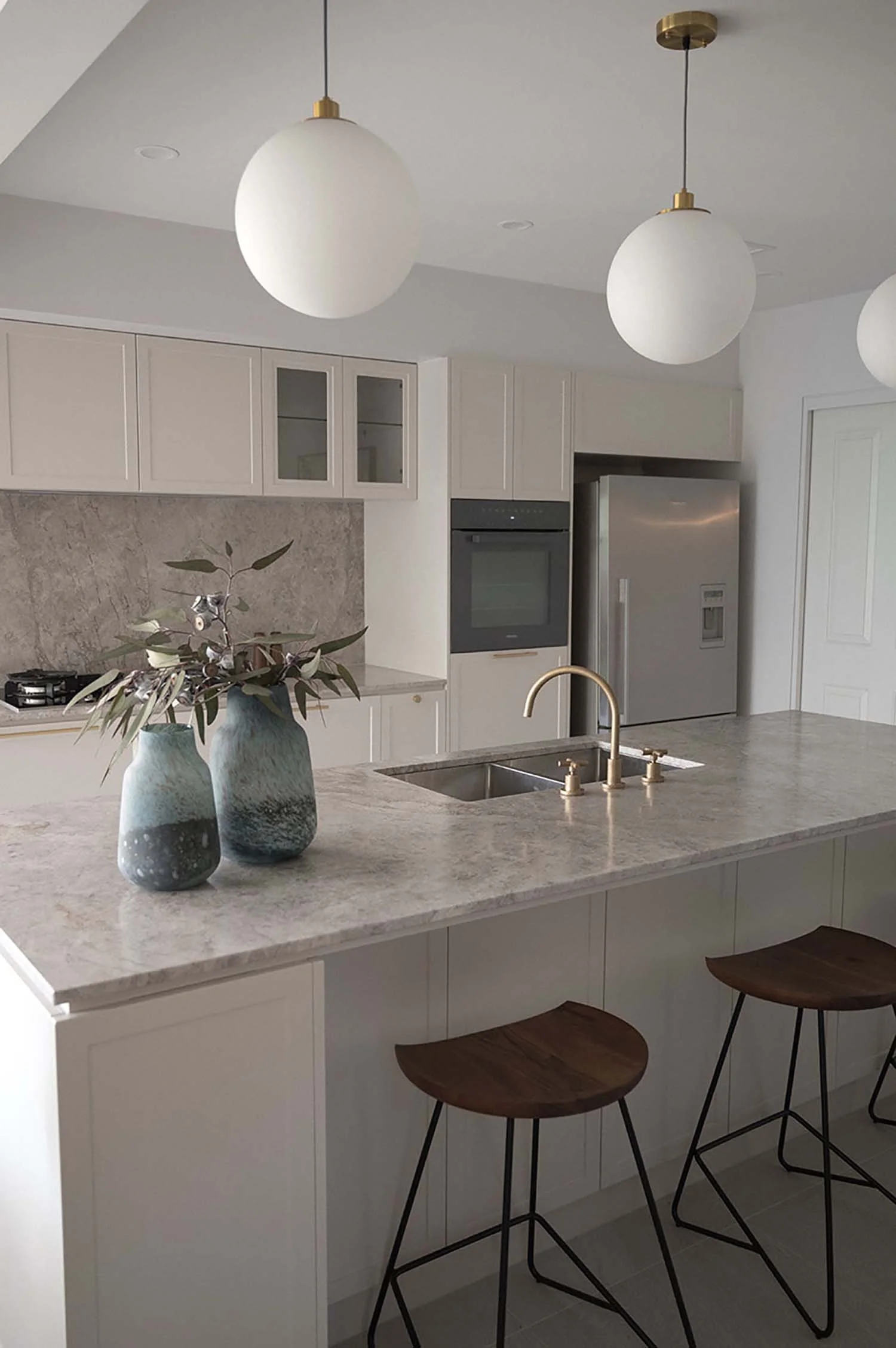 Modern kitchen with white cabinetry, marble countertop, and stainless steel refrigerator. Two wooden bar stools with metal legs are positioned at the kitchen island. The island has a double sink with a gold faucet. Decorative vases with green and whi