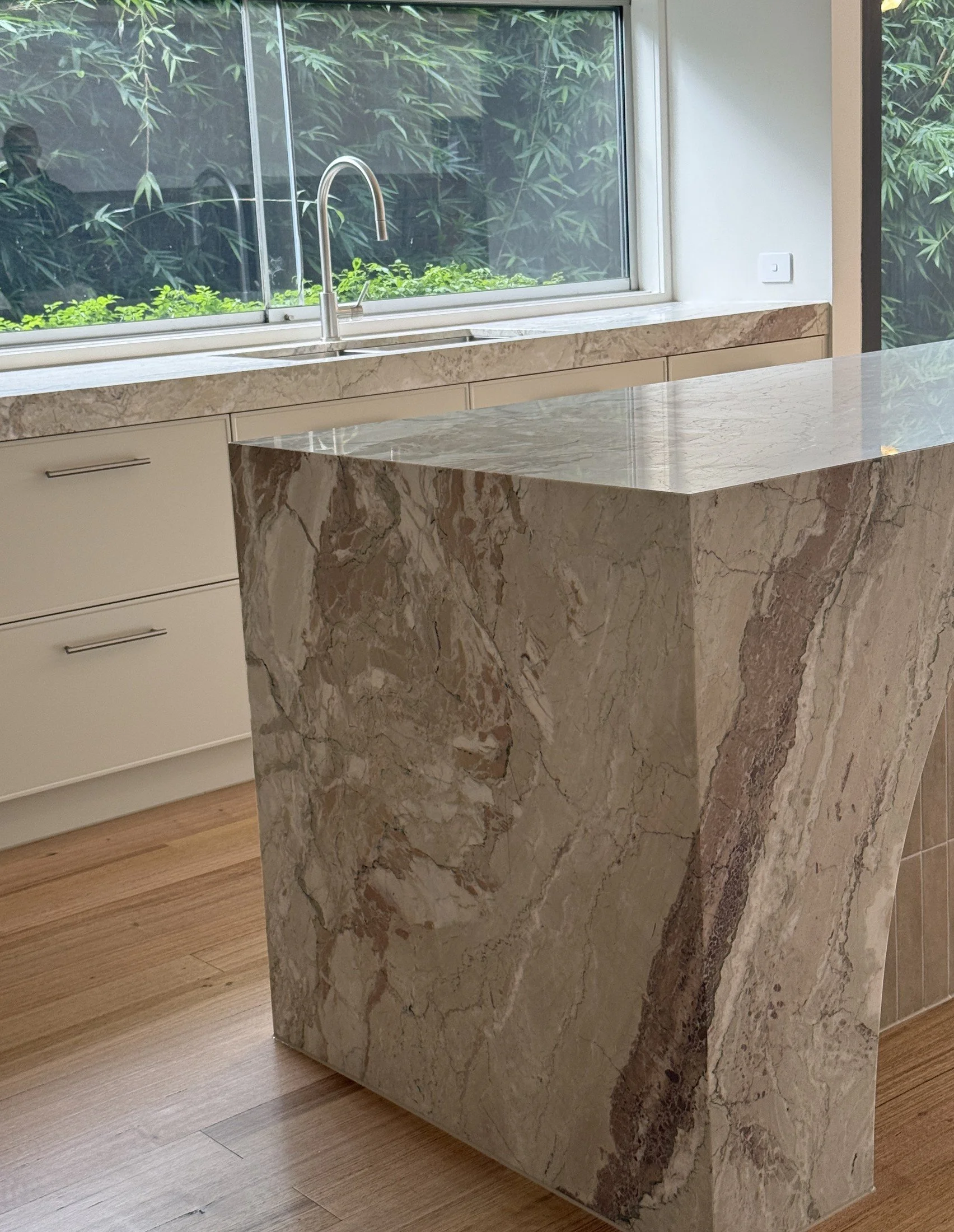 Veined stone waterfall island beside pale kitchen cabinetry and a garden-facing window.