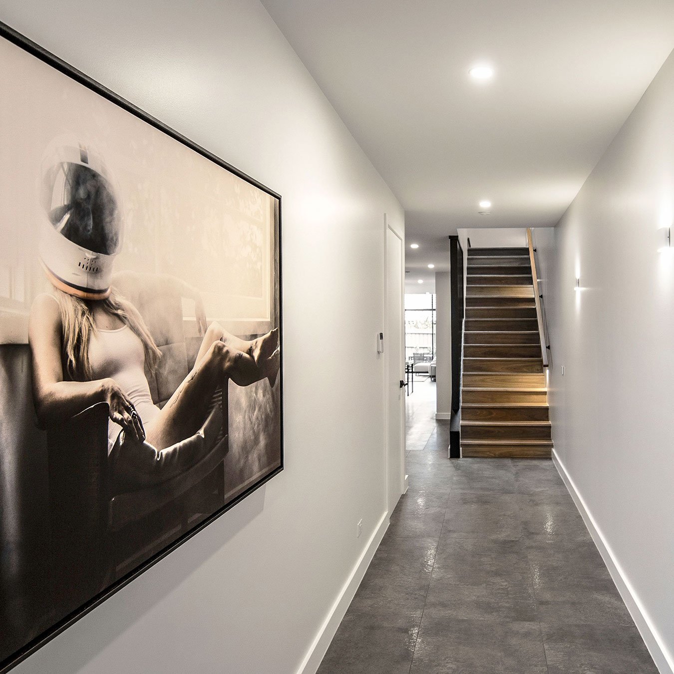 A hallway with gray tile flooring, white walls, and a staircase made of wood with black railings. There is a large framed black and white photograph of a woman wearing a swimsuit and an astronaut helmet, sitting on a chair, mounted on the left wall.