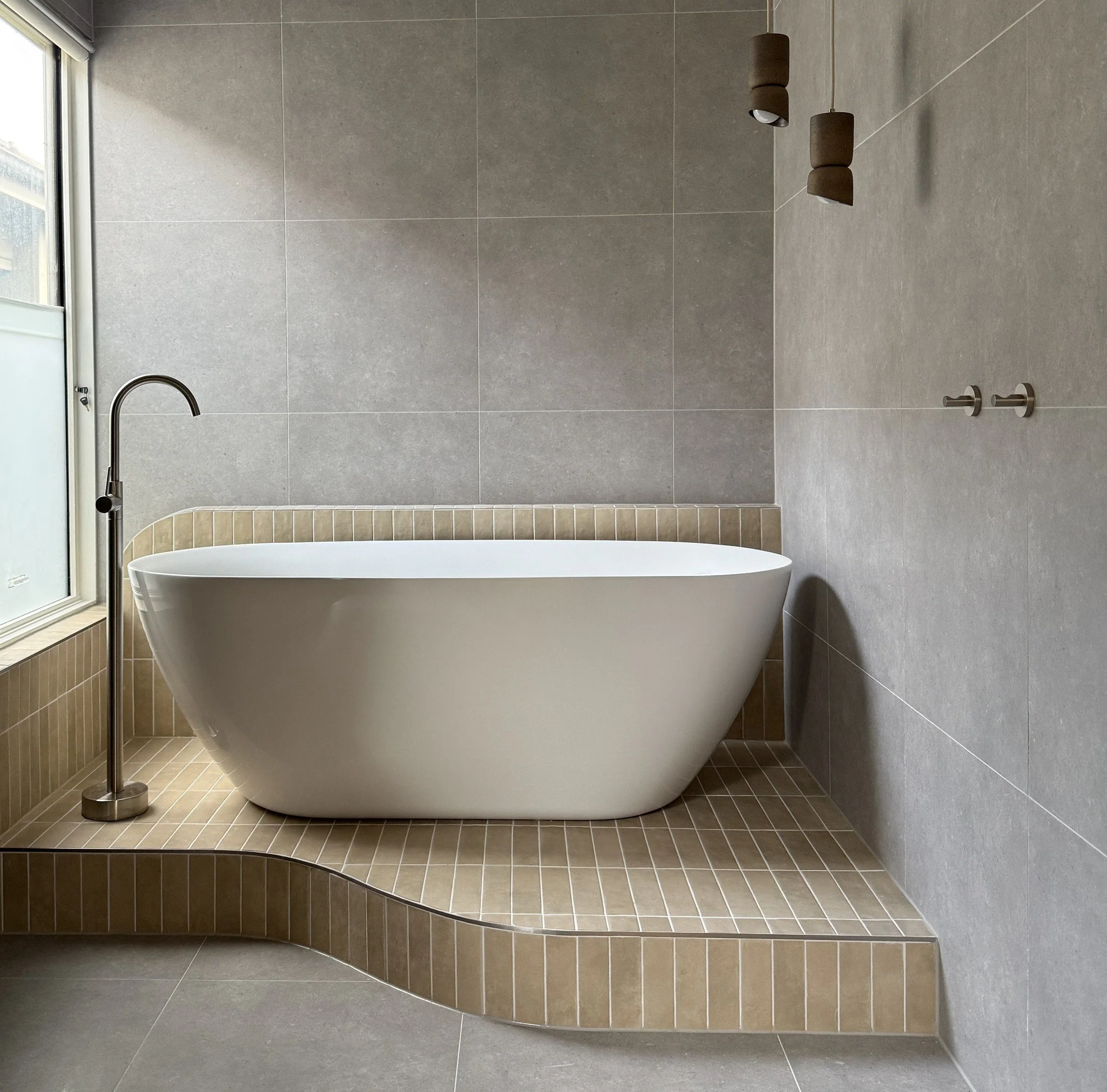 Freestanding white bath on a curved tiled plinth beneath brass wall lights beside a window.