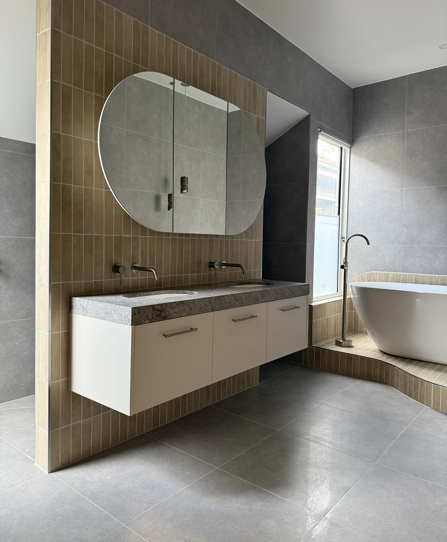 Bathroom with timber and white vanity joinery, a rounded mirror, stone flooring and a freestanding bath.