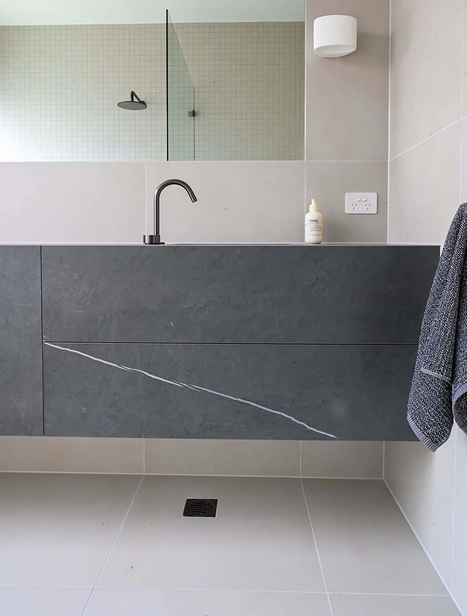 Modern bathroom with a gray vanity, a black faucet, a bottle of hand soap, a wall-mounted white light fixture, a power outlet, a towel hanging on the right, and a walk-in shower with a rain showerhead behind a glass panel.