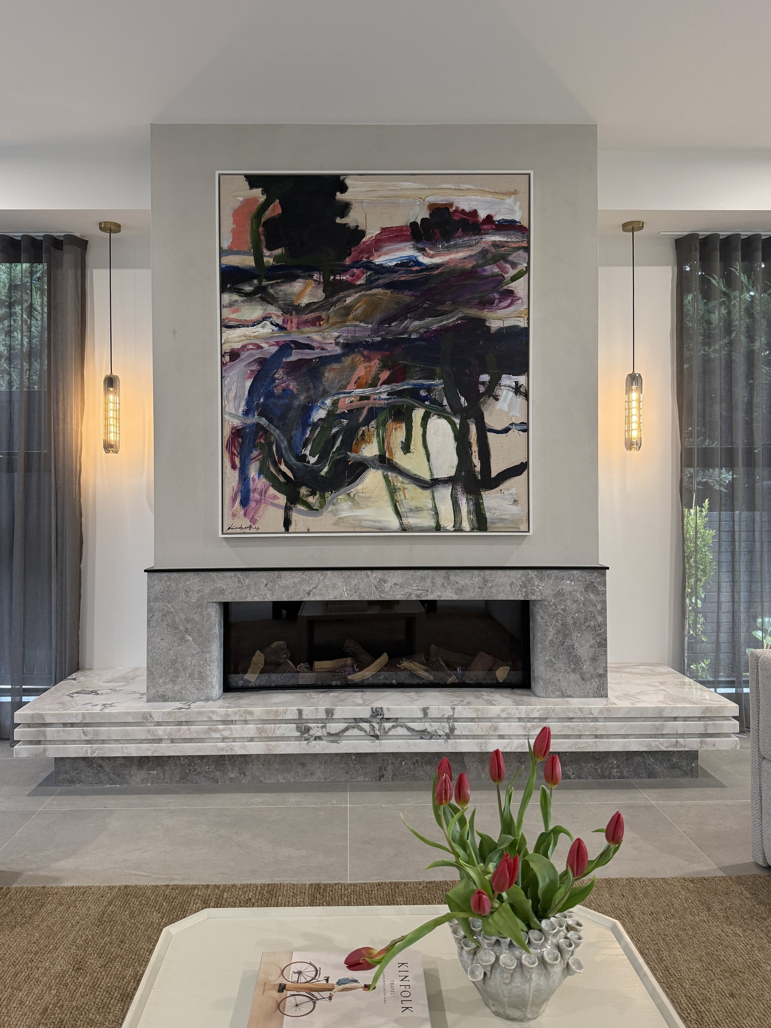Stone fireplace beneath abstract artwork, flanked by pendant lights and dark sheer curtains.