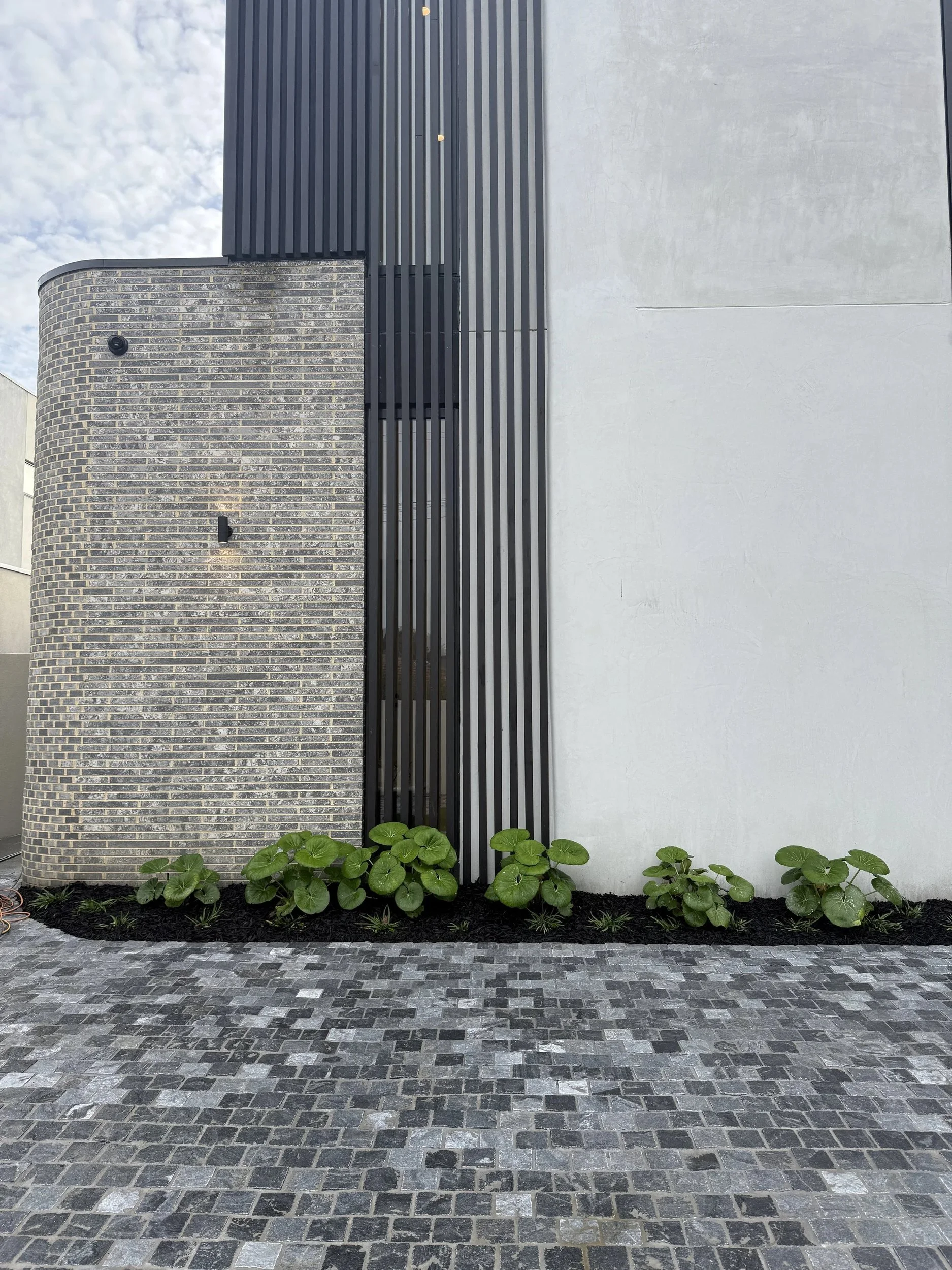 Exterior detail with textured grey masonry, dark vertical cladding, low planting and stone paving.