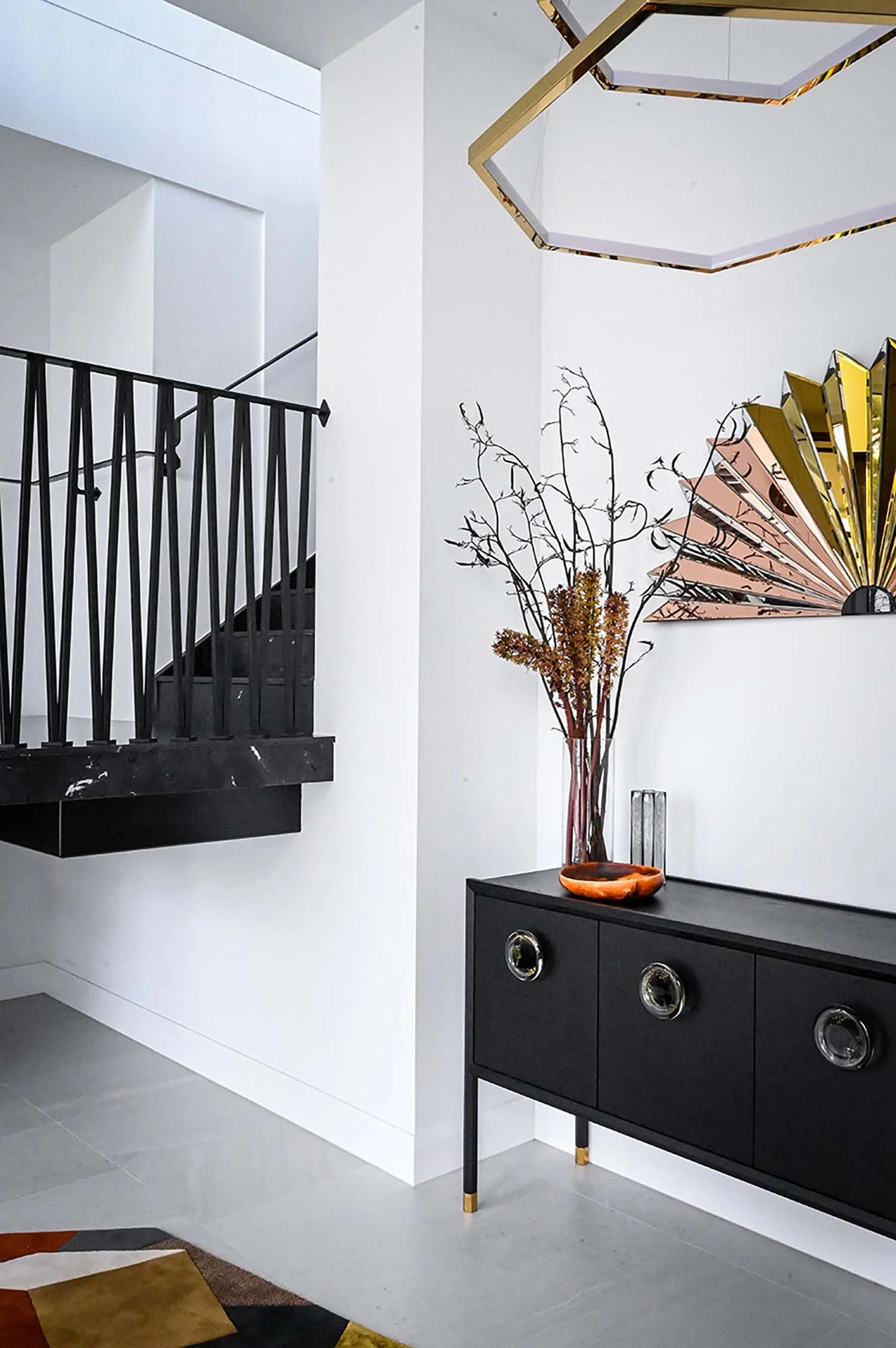 Modern interior with a black sideboard, decorative vase with dried branches, an orange bowl, abstract wall art, and a staircase with black railing.