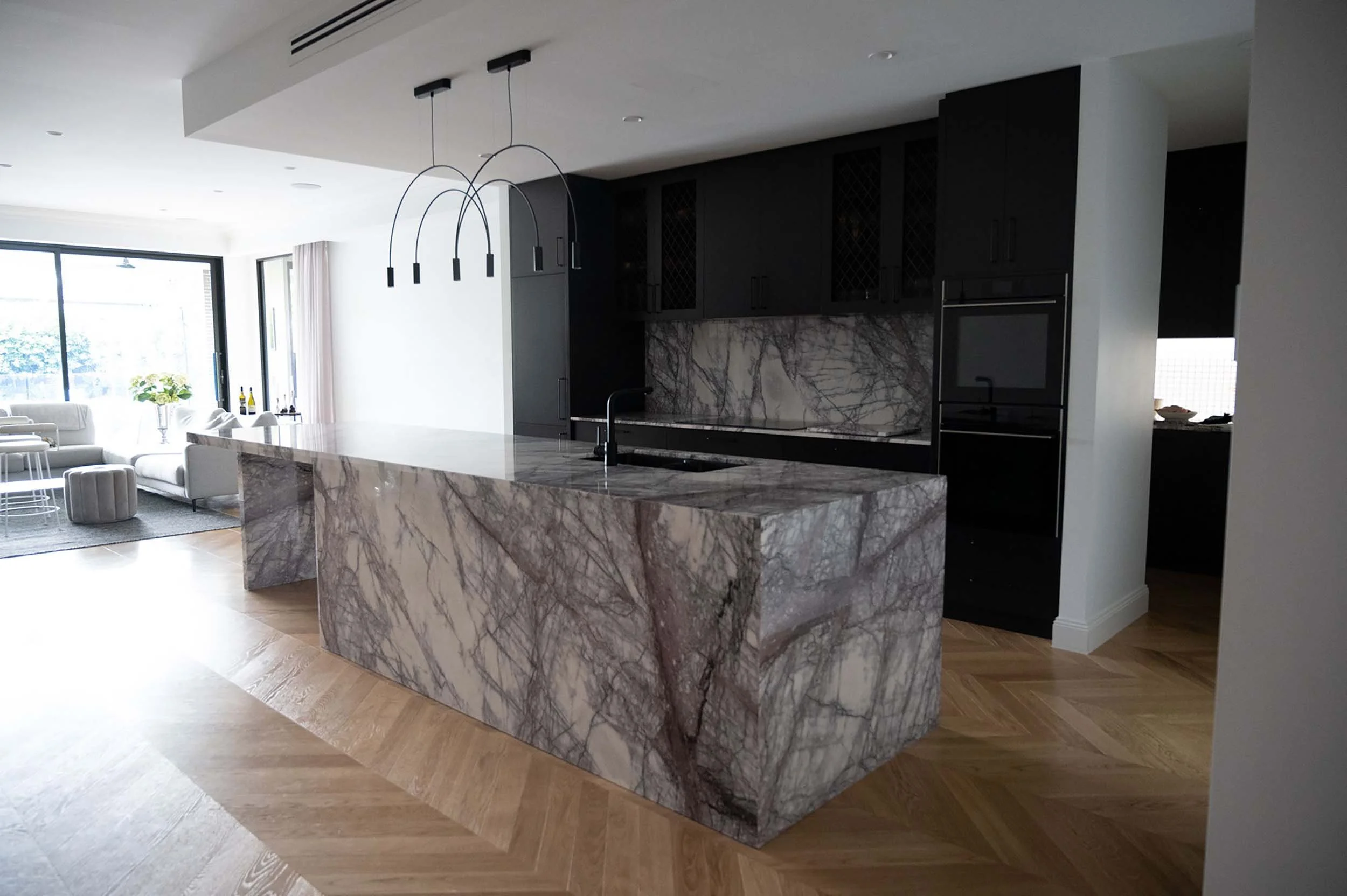 Modern kitchen with a marble island, black cabinets, pendant lighting, and an open living area with large windows.