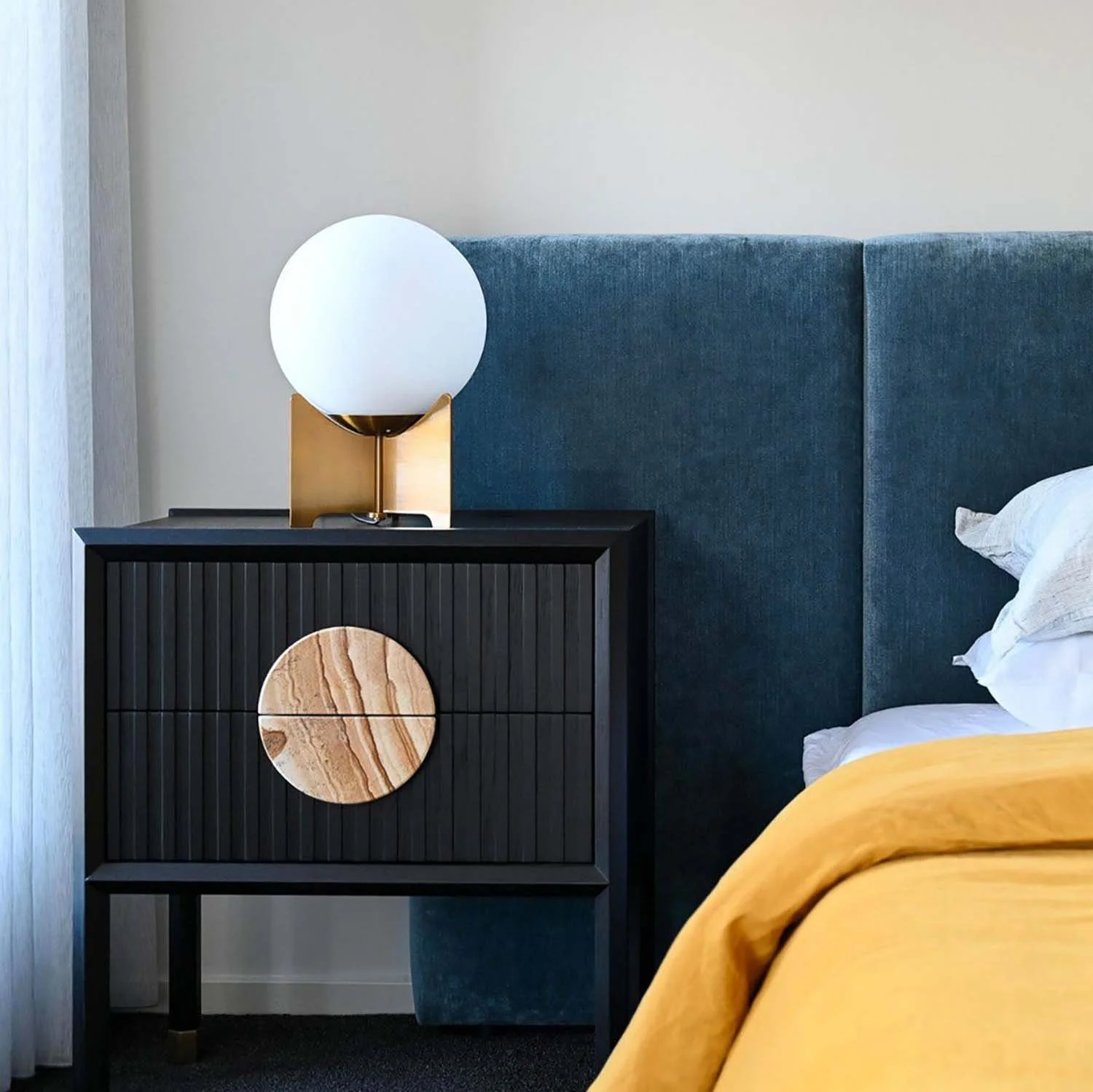 A bedroom corner featuring a black nightstand with a wooden circular accent and vertical grooves, a modern lamp with a spherical white shade and a gold base, a blue fabric headboard, a bed with white sheets and a yellow blanket, and light-colored cur
