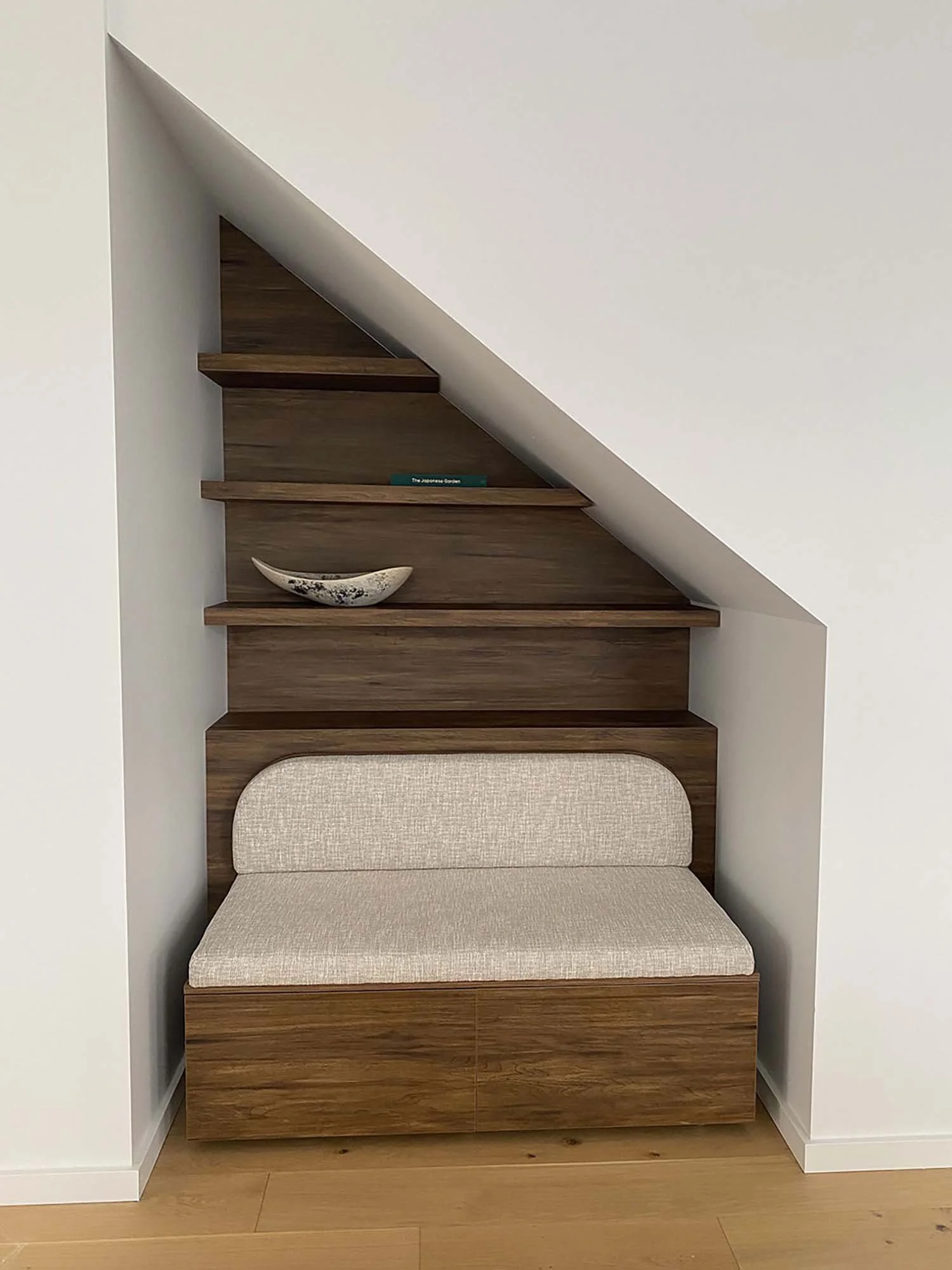 A modern staircase with wooden steps built into a wall, featuring a built-in shelf with a green book and a decorative bowl, and a beige cushioned seat at the bottom.