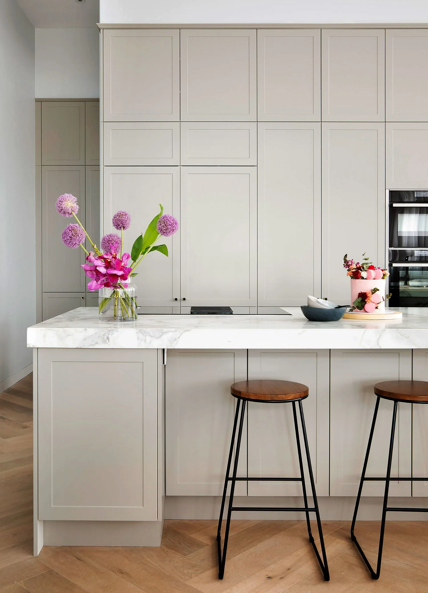 A modern kitchen with a marble countertop and beige cabinets, decorated with pink flowers in a glass vase and pink macarons on a round tray.
