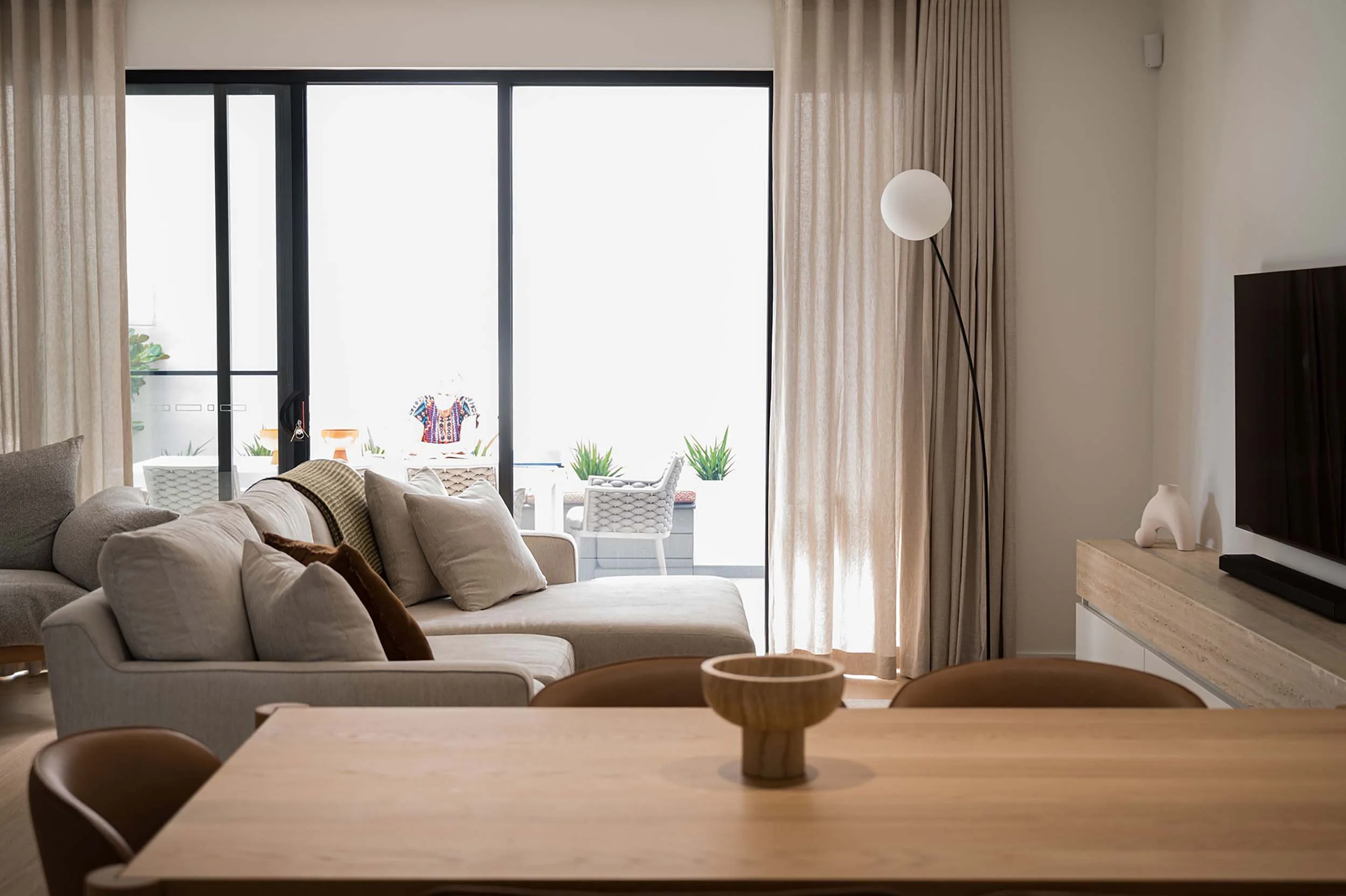 Living room with beige sofa, chairs, and beige curtains, open to a balcony with outdoor chairs, plants, and a colorful shirt hanging outside.