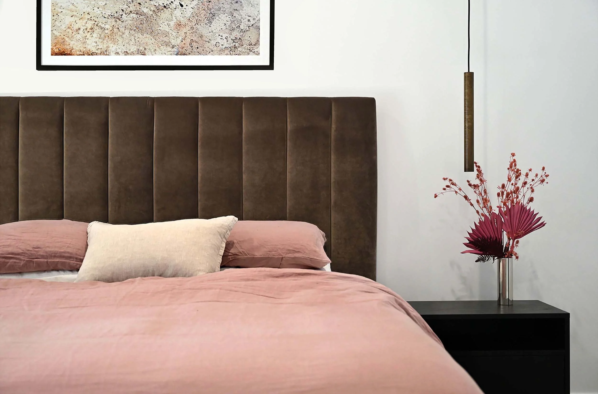 A bedroom with a bed dressed in pink bedding and beige pillows, a dark brown velvet headboard, a black nightstand with a tall metallic vase holding red and pink flowers, a framed artwork above the bed, and a hanging pendant light with a brass finish.