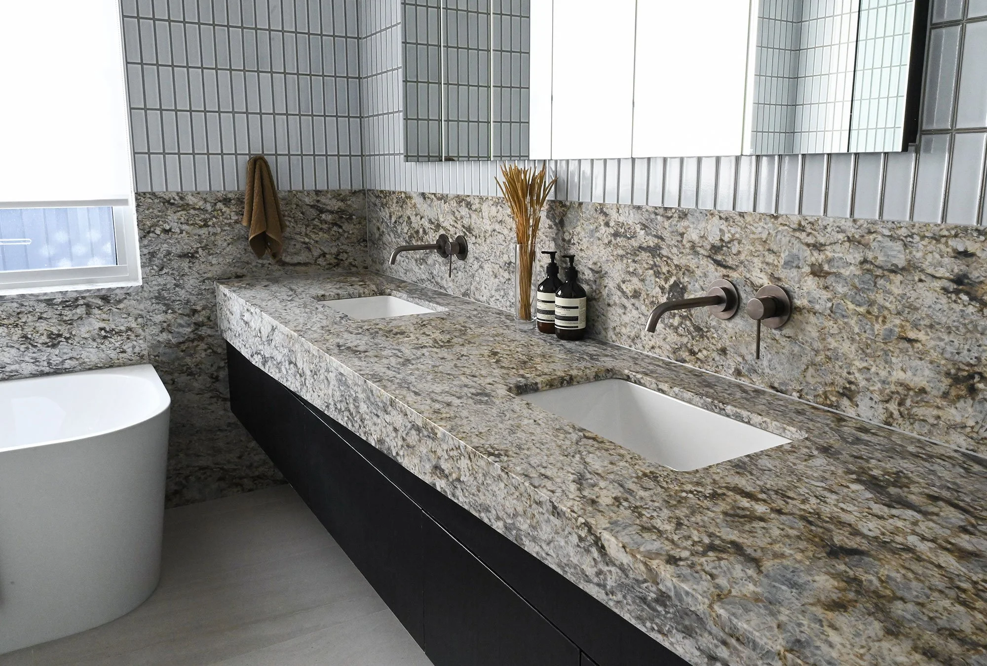 Bathroom with a granite countertop, two sinks, minimalistic decor, brown towels, bottles, and a tall vase with dried grass, white tiled wall, and a large mirror.