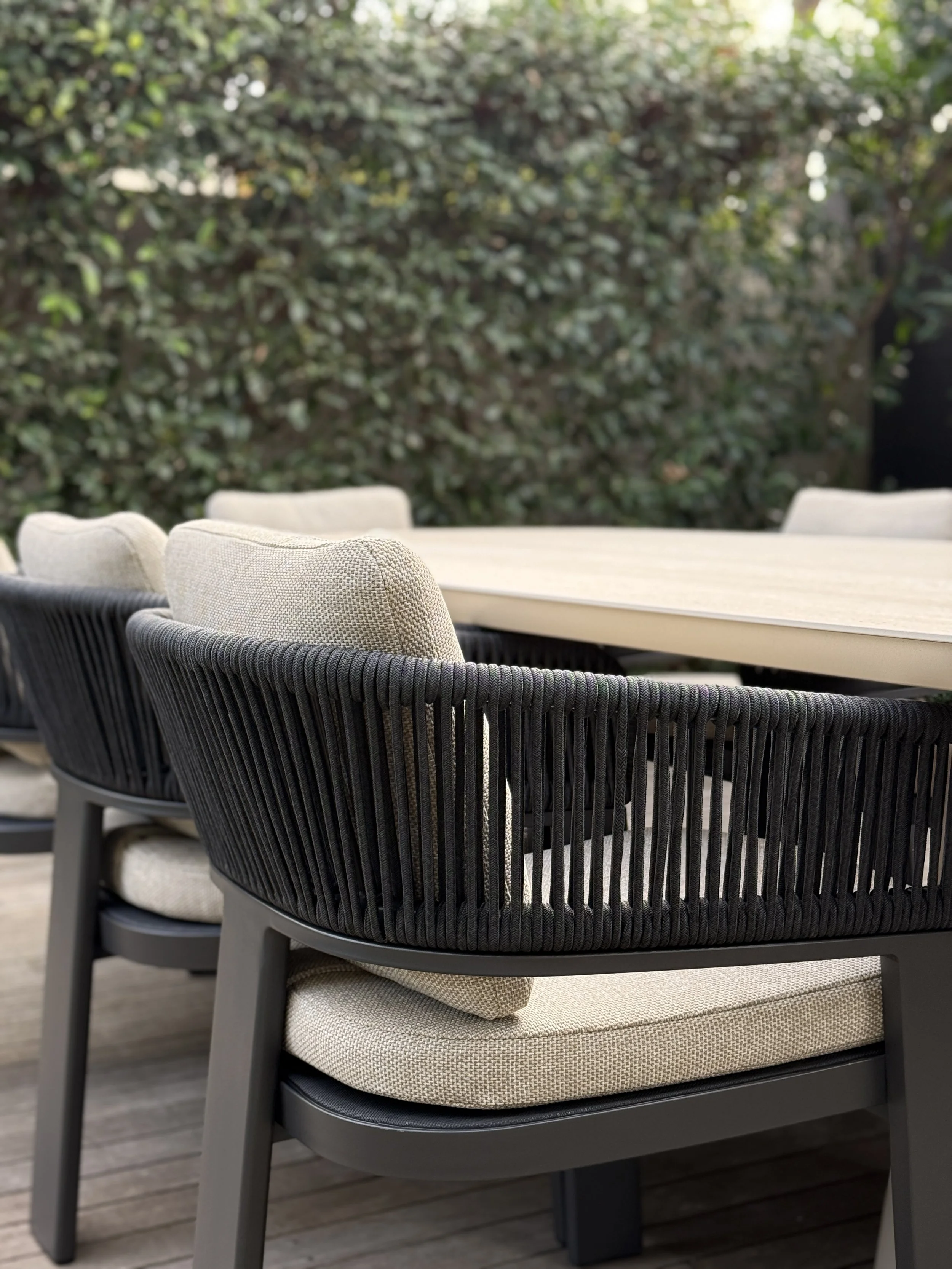 Outdoor patio with beige cushioned chairs and a long beige table against a backdrop of green bushes.