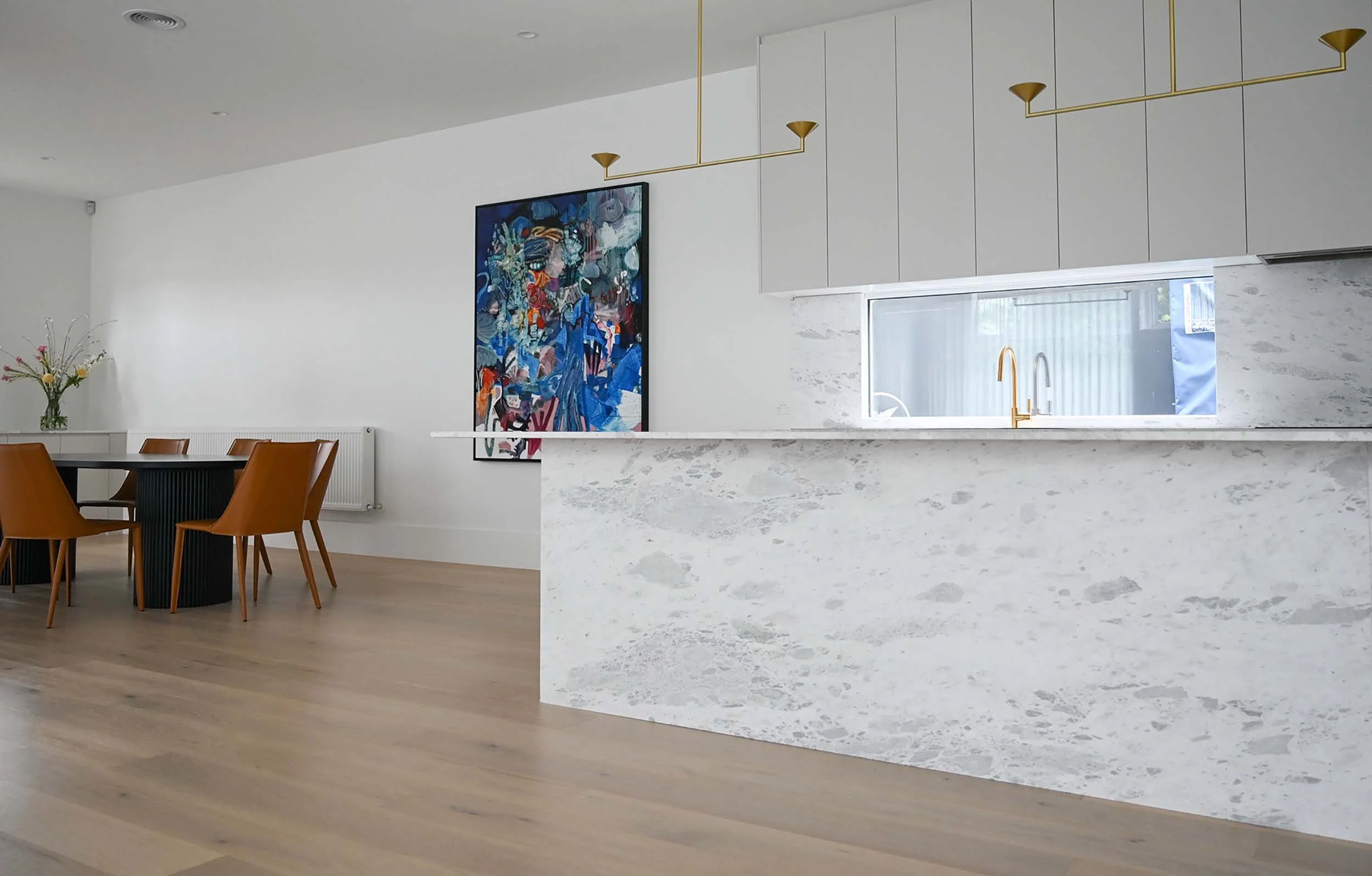 Modern kitchen with a marble island, white cabinets, a window with a gold faucet, and a dining area with brown chairs and a vase of flowers.