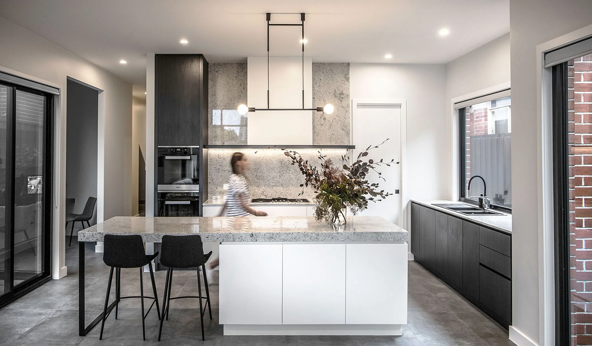 Modern kitchen with large island, gray cabinets, and a woman walking past. Bright space with large windows and black pendant lights.