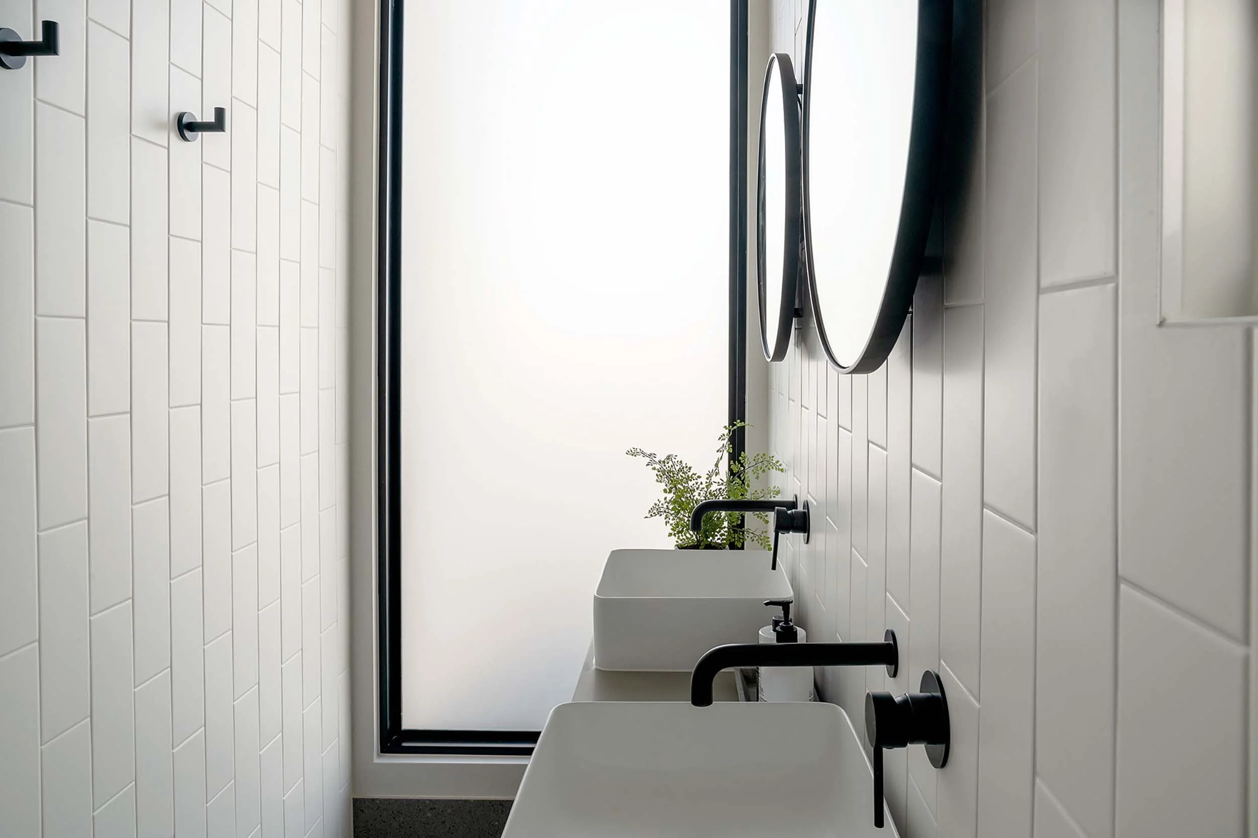 Modern bathroom with white tiled walls, black fixtures, a large window letting in natural light, and a small green plant on the windowsill.