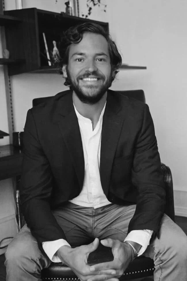 Man sitting in chair, smiling, wearing a suit jacket and shirt.