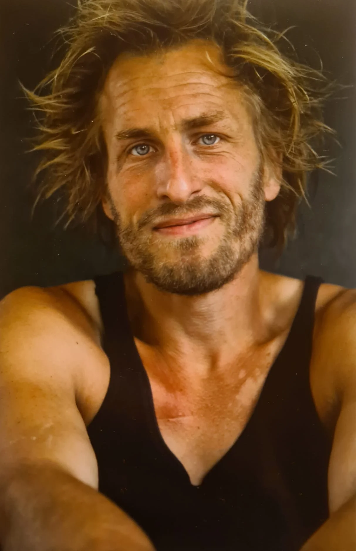 A portrait of a man with messy hair, beard, and a black sleeveless shirt, smiling slightly, with a dark background.