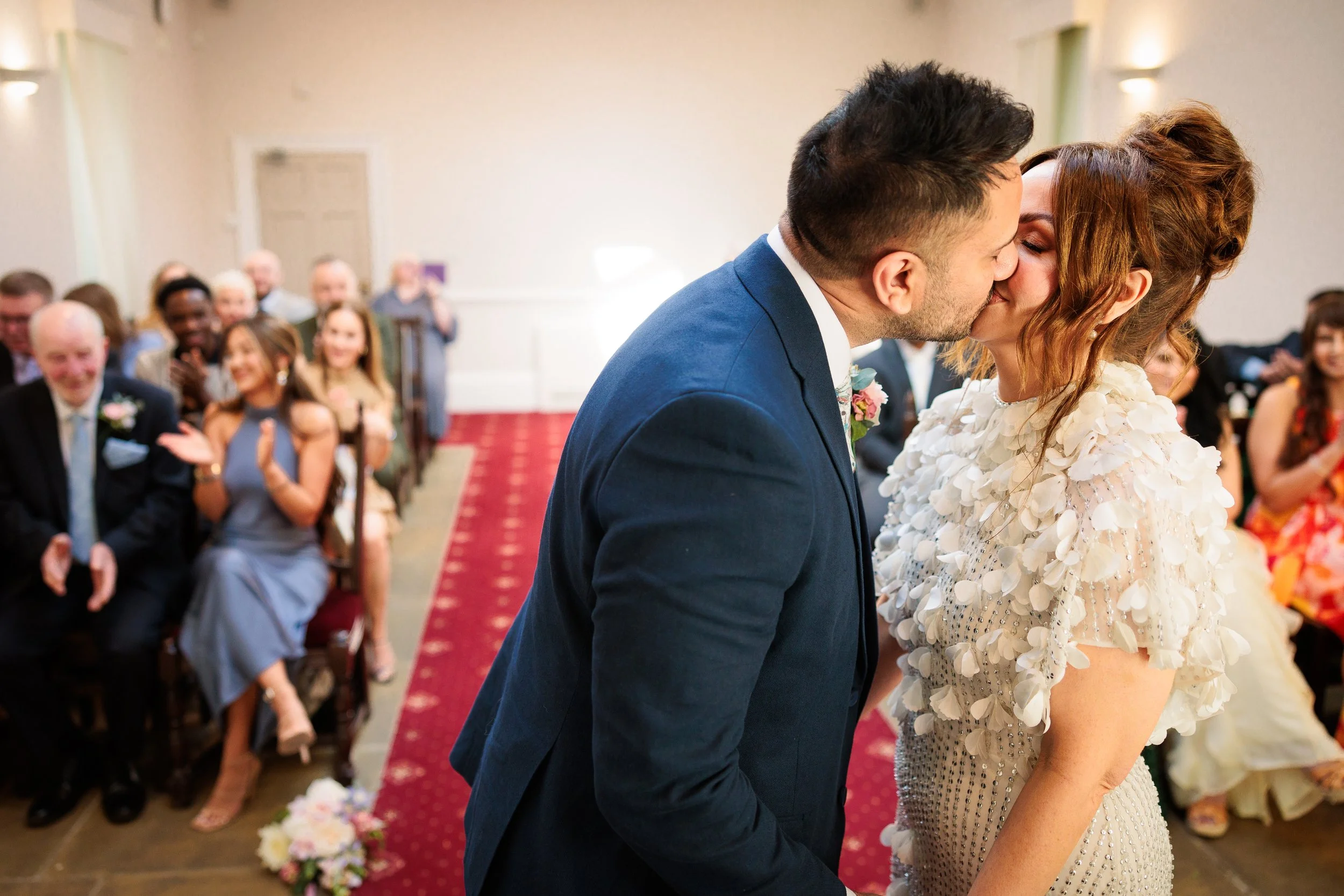 A couple is kissing at their wedding ceremony surrounded by seated guests who are clapping and smiling.
