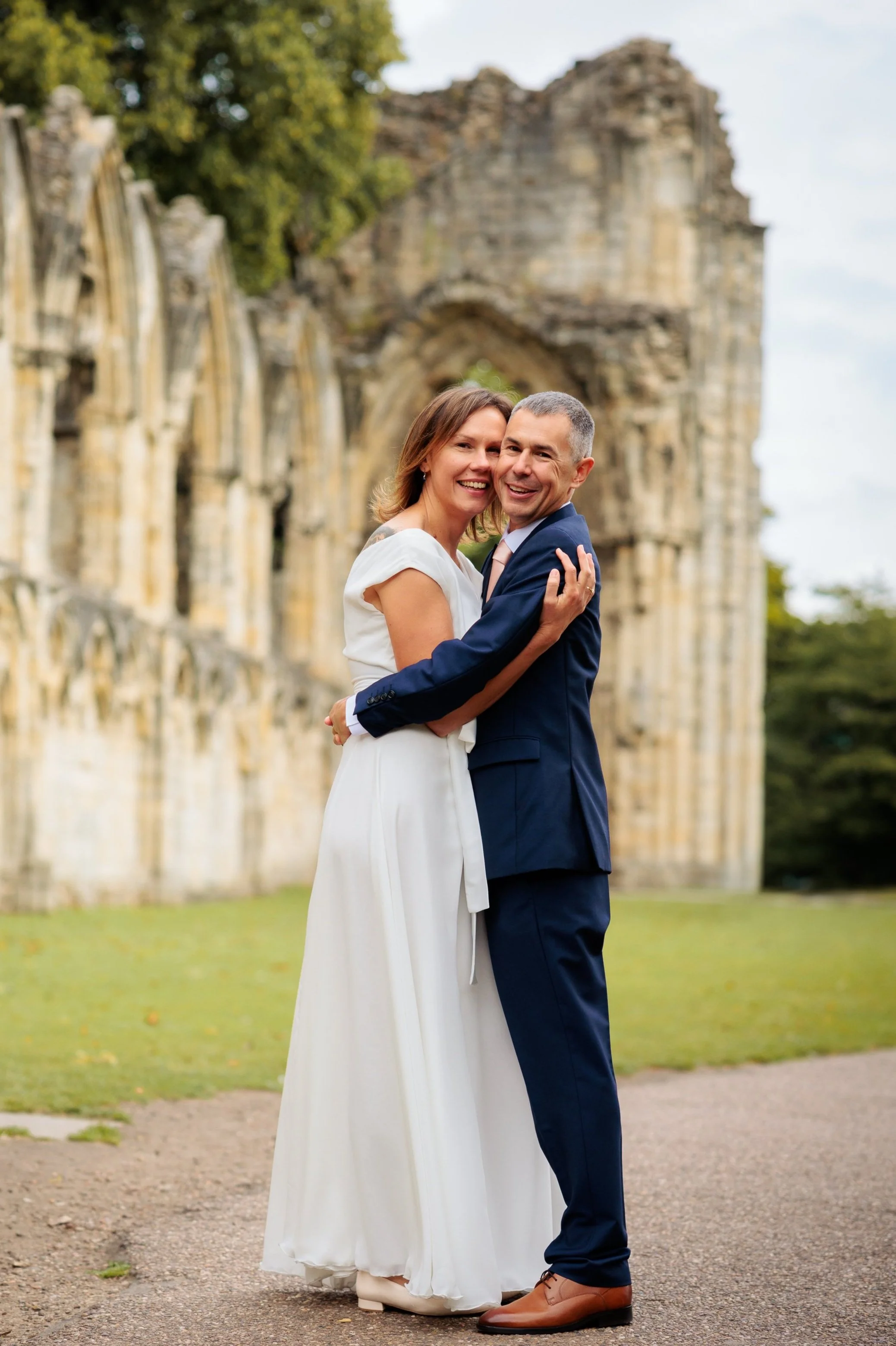 A happy couple in wedding attire embracing outdoors in front of ancient ruins.
