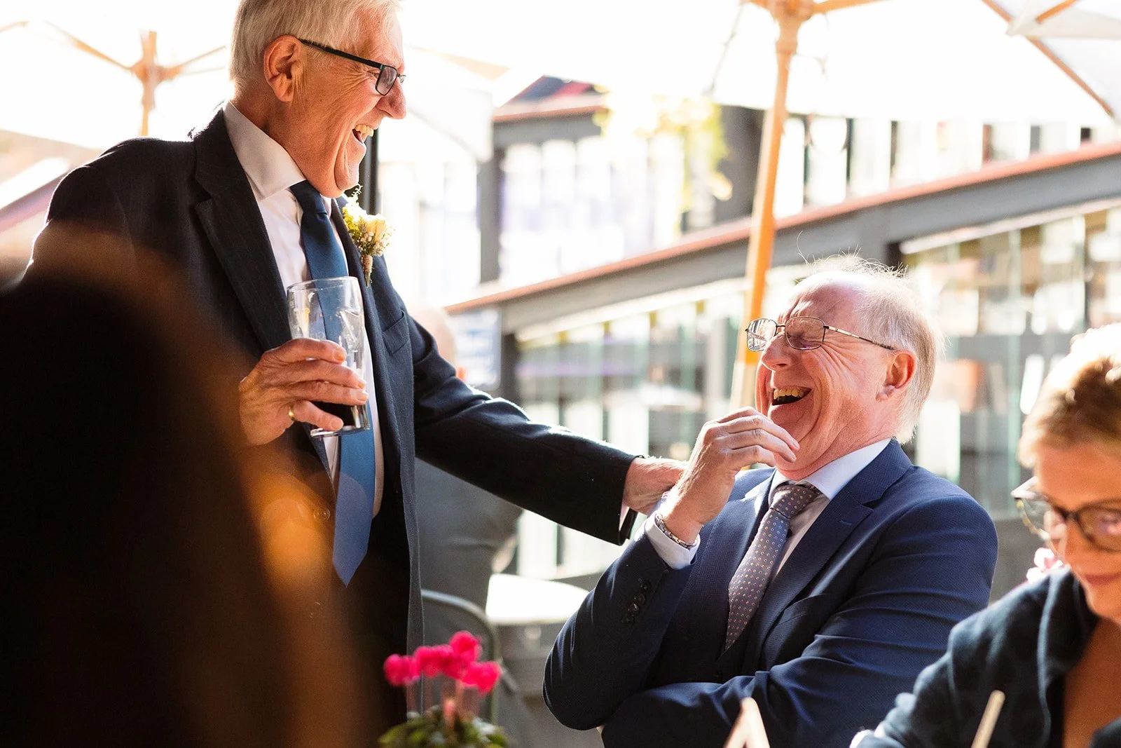 Two elderly men in suits and glasses are sharing a joyful moment, laughing and smiling at each other in an outdoor setting with umbrellas.