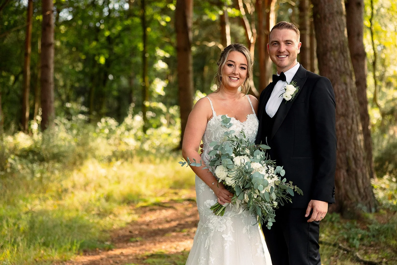 A bride and groom smiling and standing together in a wooded outdoor setting, dressed in wedding attire. The bride is holding a large bouquet of white flowers and greenery, wearing a white lace wedding gown. The groom is wearing a black tuxedo with a 