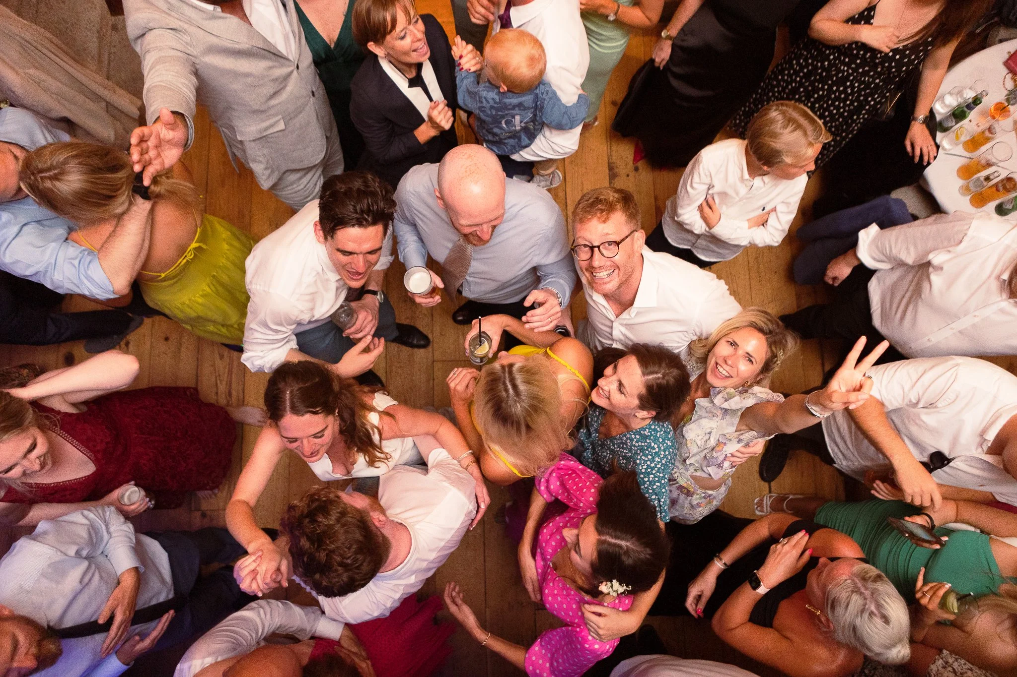 Partygoers at a social gathering, looking up at the camera, with some smiling and making peace signs, on a wooden floor with drinks and tables visible.