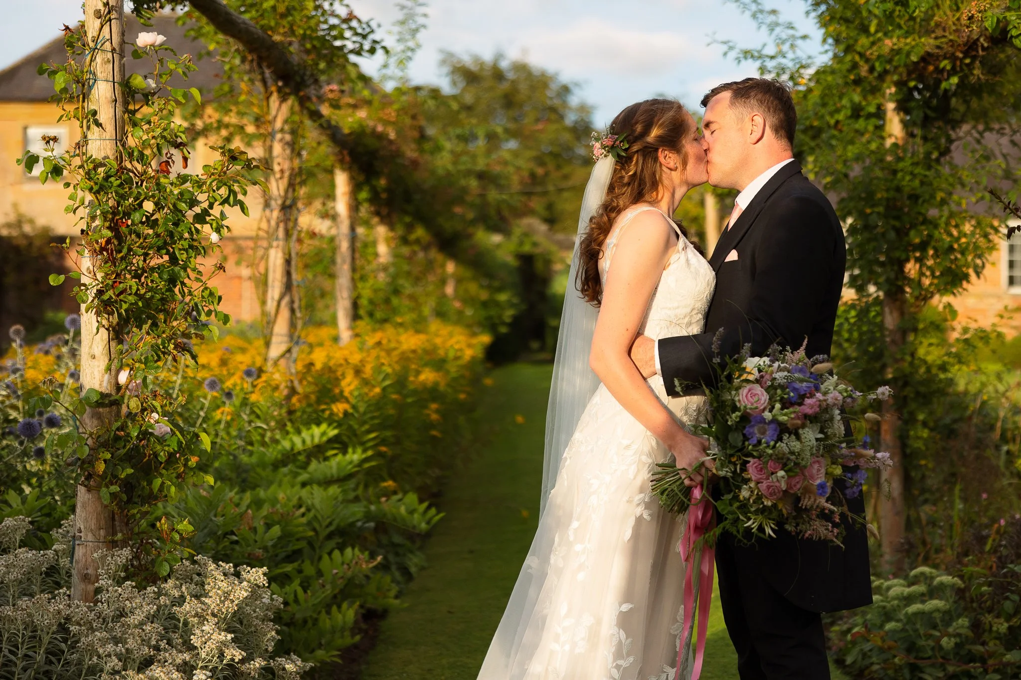 A bride and groom sharing a kiss in a garden, with the bride holding a bouquet and dressed in a white wedding gown, while the groom wears a black tuxedo. The garden has lush green plants and colorful flowers.