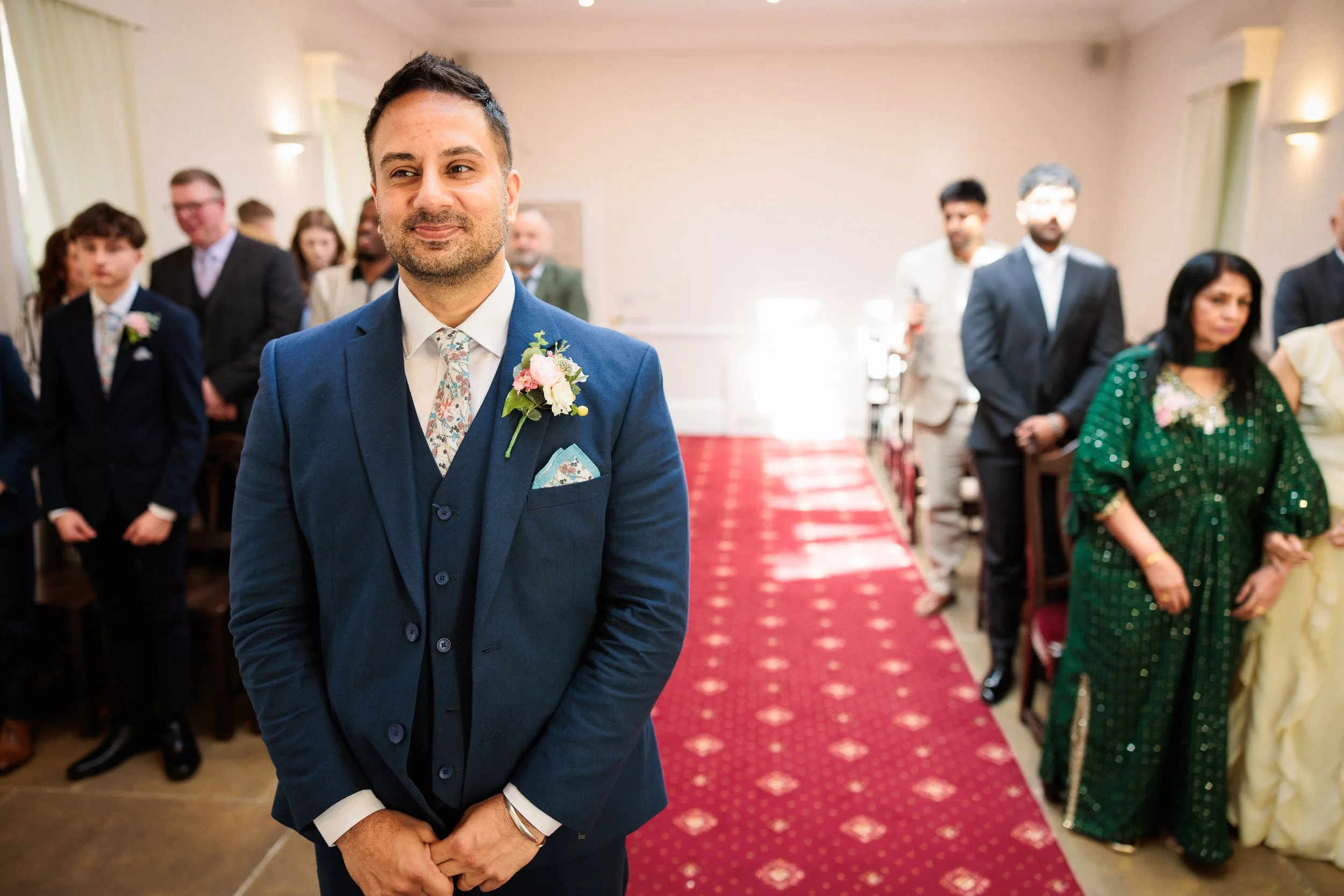 A groom in a blue suit with a floral boutonniere stands at a wedding ceremony, surrounded by guests in formal attire, inside a decorated venue with a red carpet aisle.