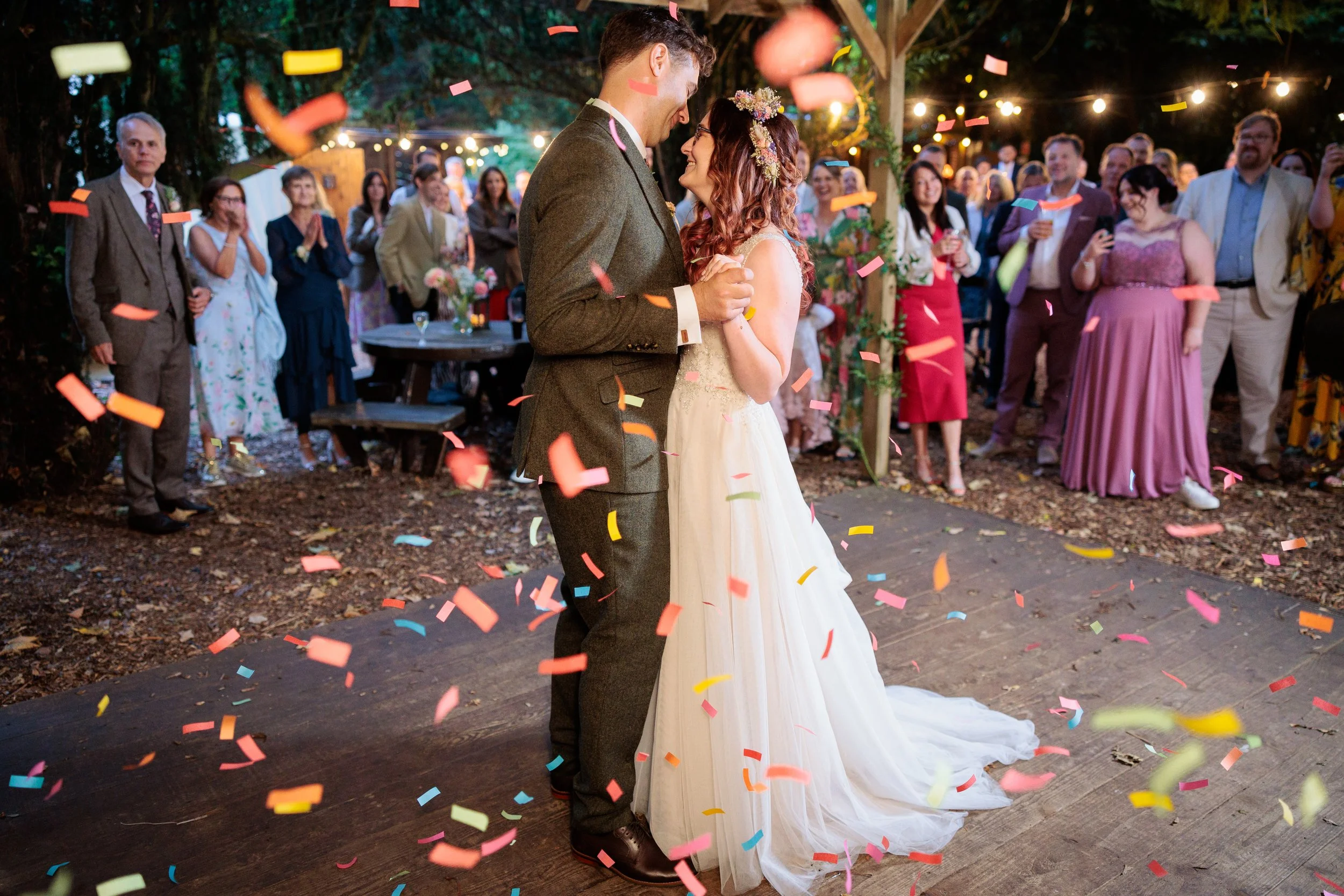 A bride and groom dancing under string lights at their outdoor wedding reception, surrounded by guests and colorful confetti.